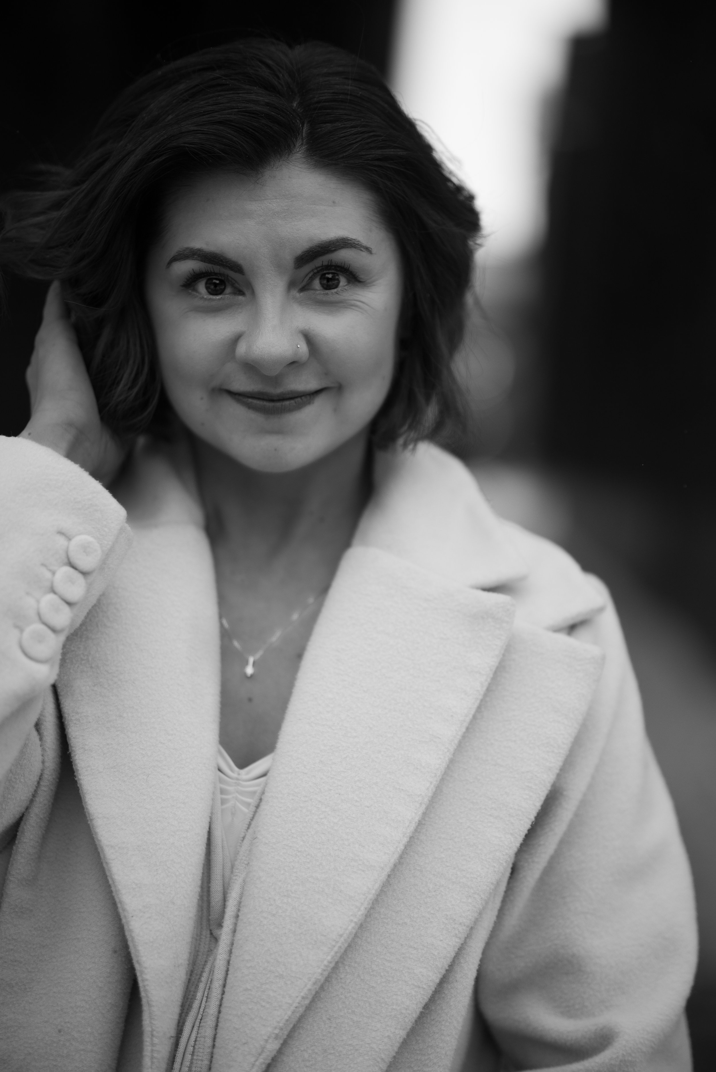 A woman with shoulder-length hair, smiling, wearing a light-colored coat and a necklace, touching her hair with her left hand.