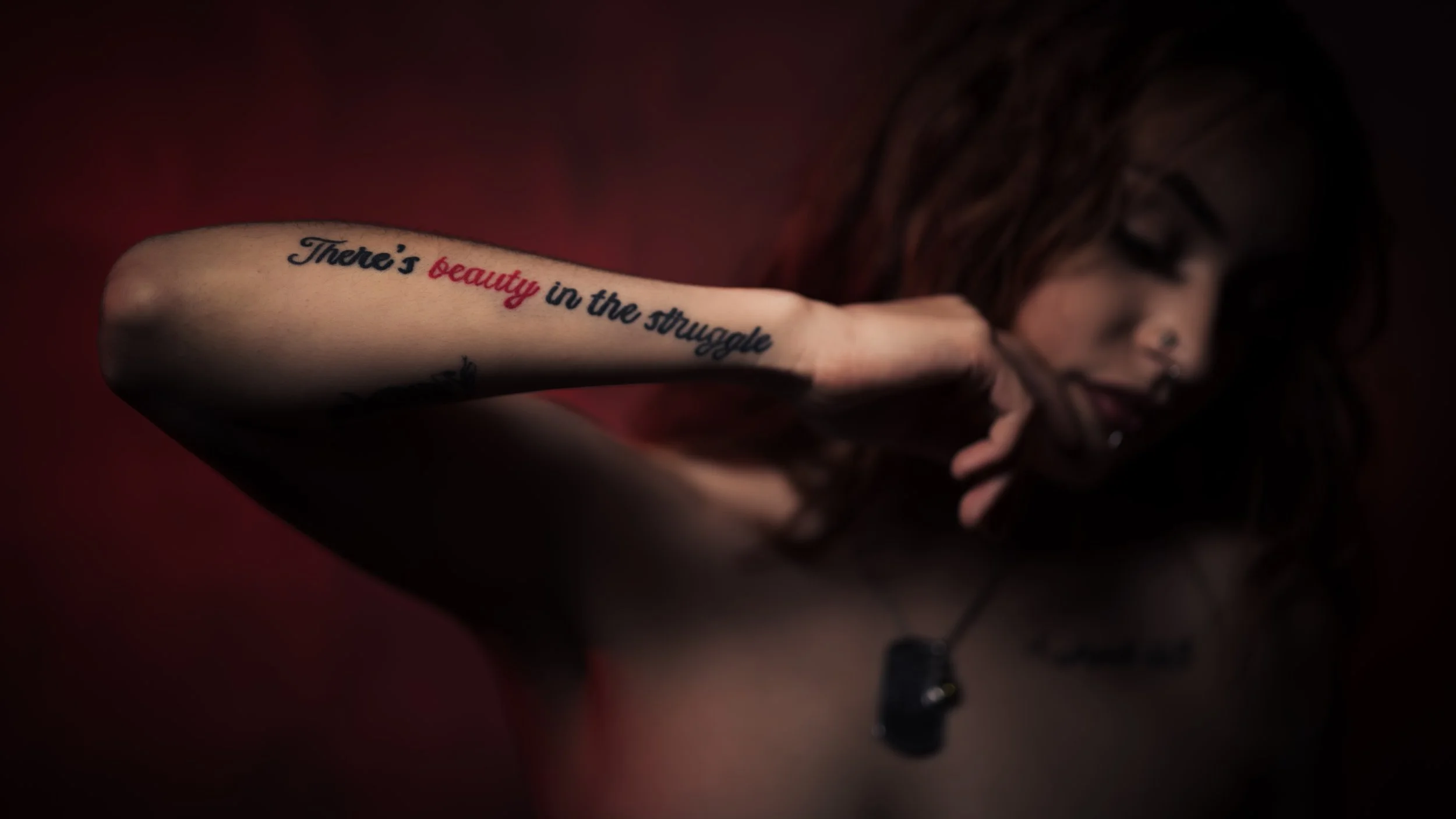 A woman with tattoos, including a quote on her arm that reads 'There's beauty in the struggle', touching her face with her hand, with a dark, blurred background.