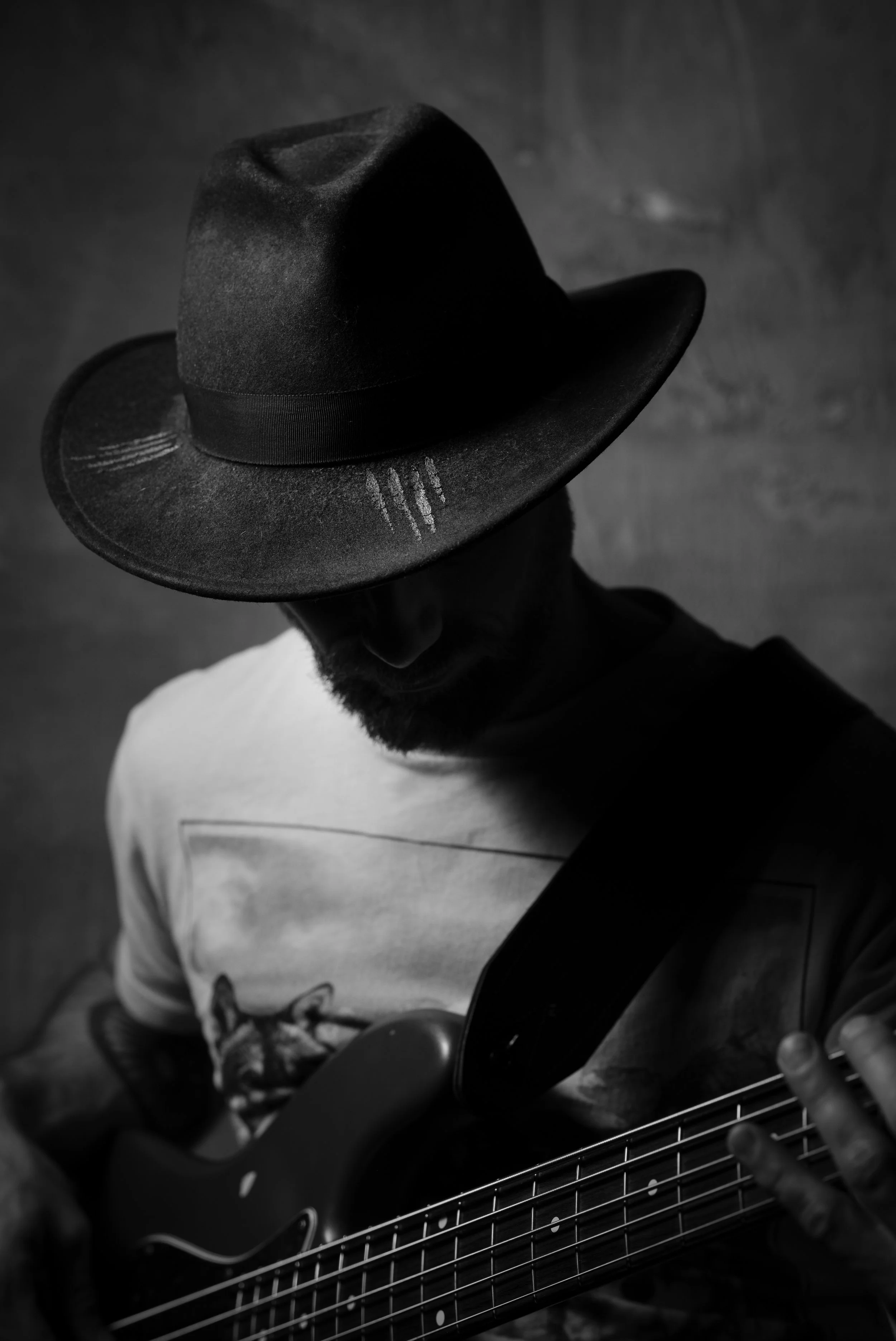 A man with a beard wearing a wide-brimmed hat and a t-shirt playing an electric guitar in a dimly lit setting.