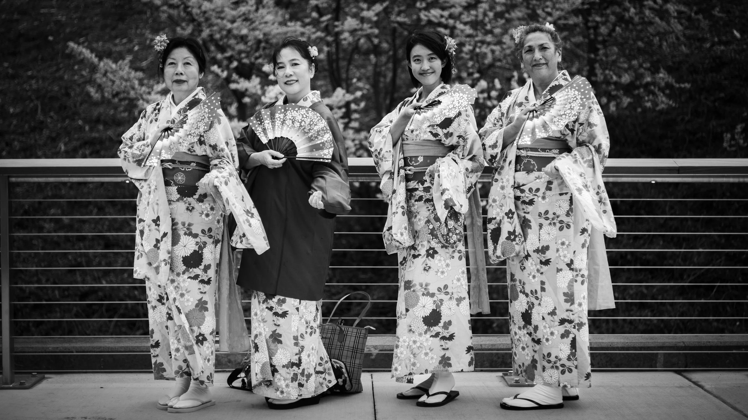 Four women wearing traditional Japanese kimonos, holding fans, standing on a wooden platform with a railing, with trees in the background.