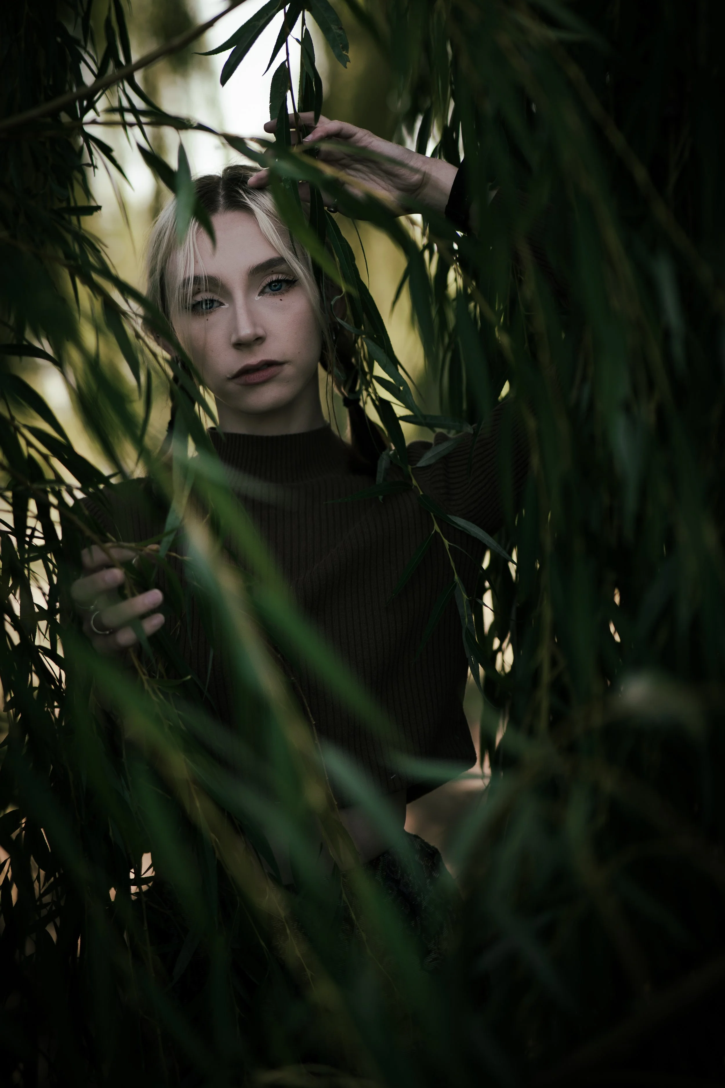 A woman with blonde hair and blue eyes posing among green plants, wearing a black top.