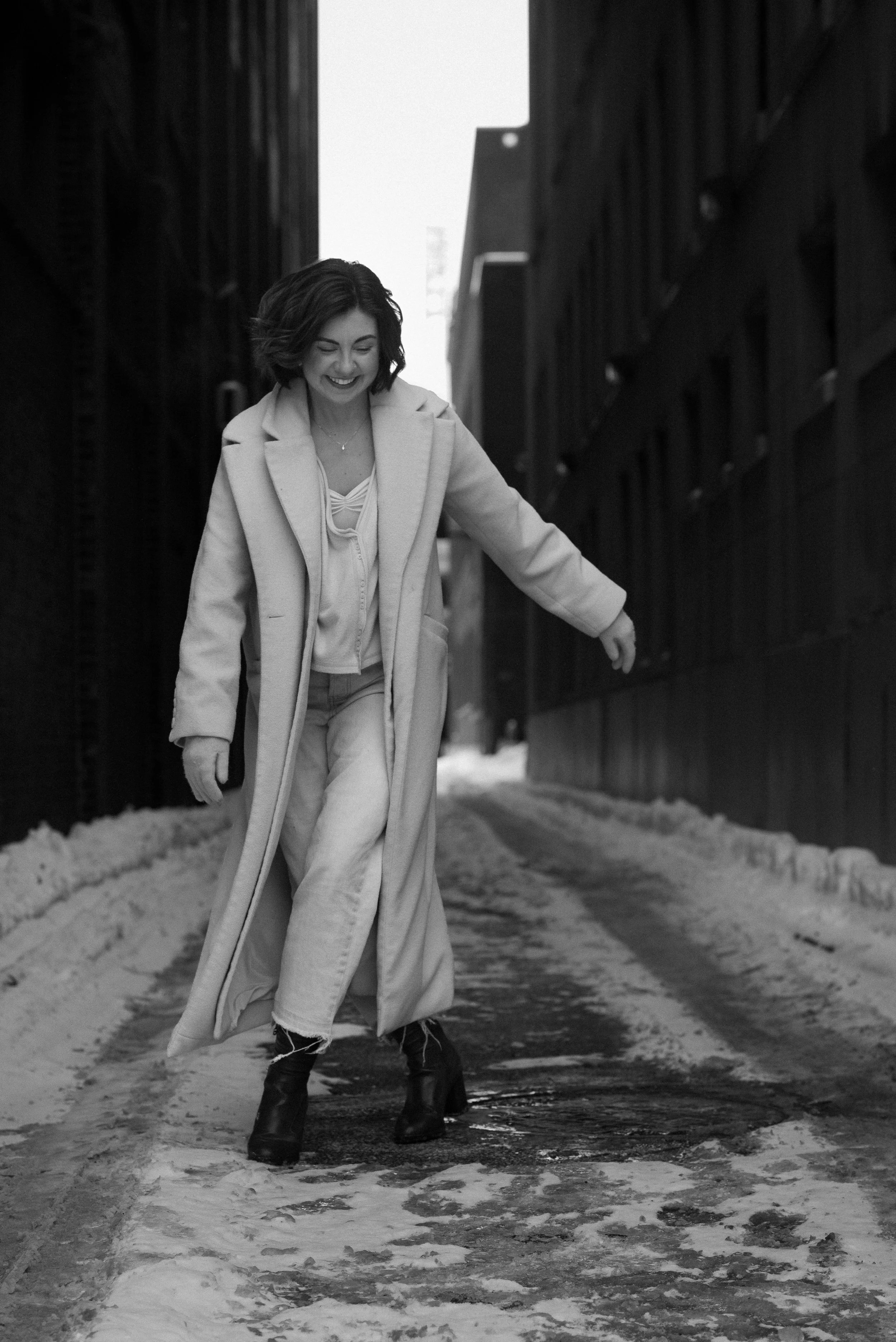 A woman walking on a snow-covered alleyway between tall buildings, smiling and looking down, wearing a long coat, jeans, and boots.