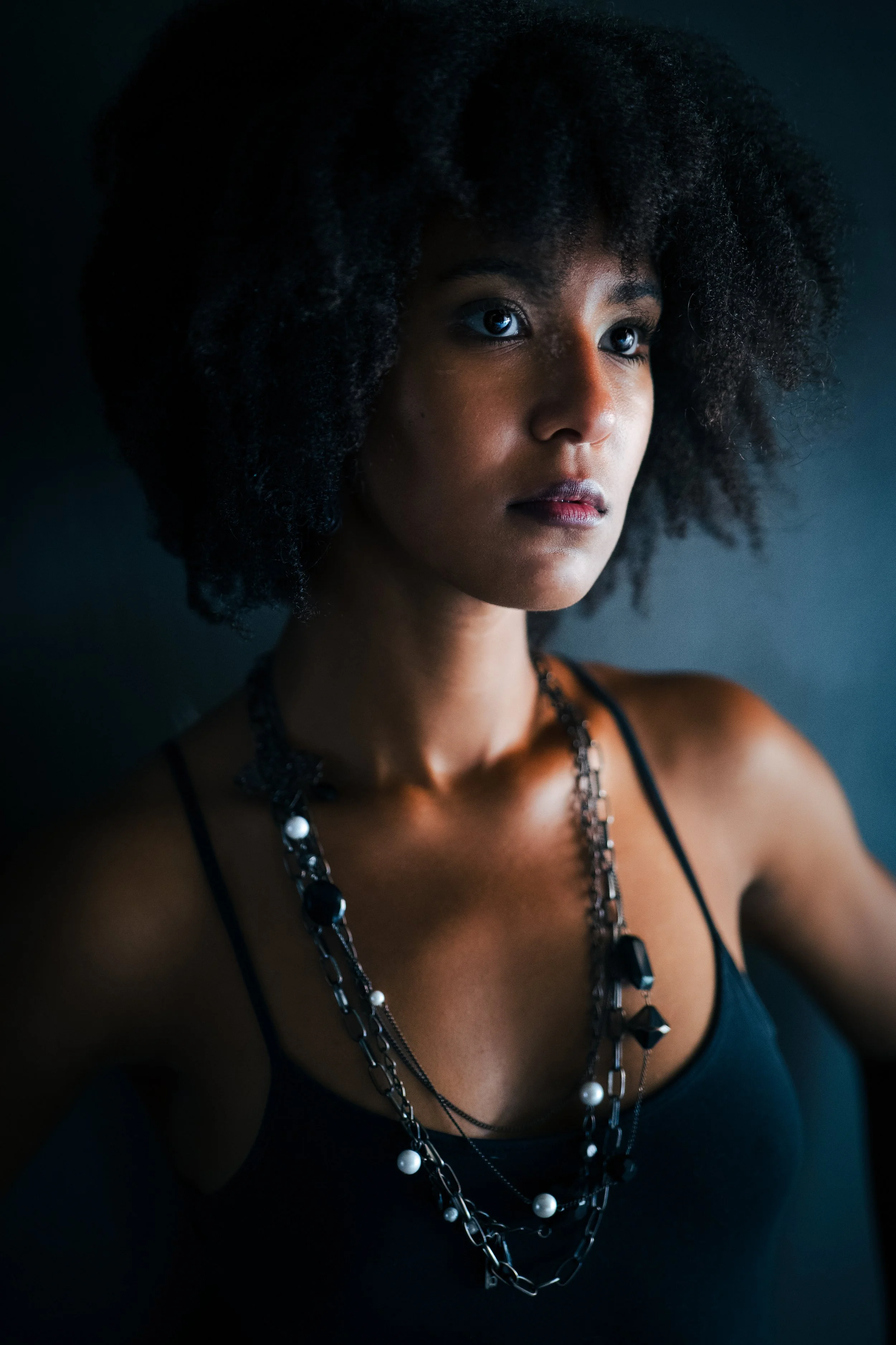 Portrait of a woman with dark, curly hair wearing a black tank top and layered necklaces, looking thoughtfully to the side against a dark background.