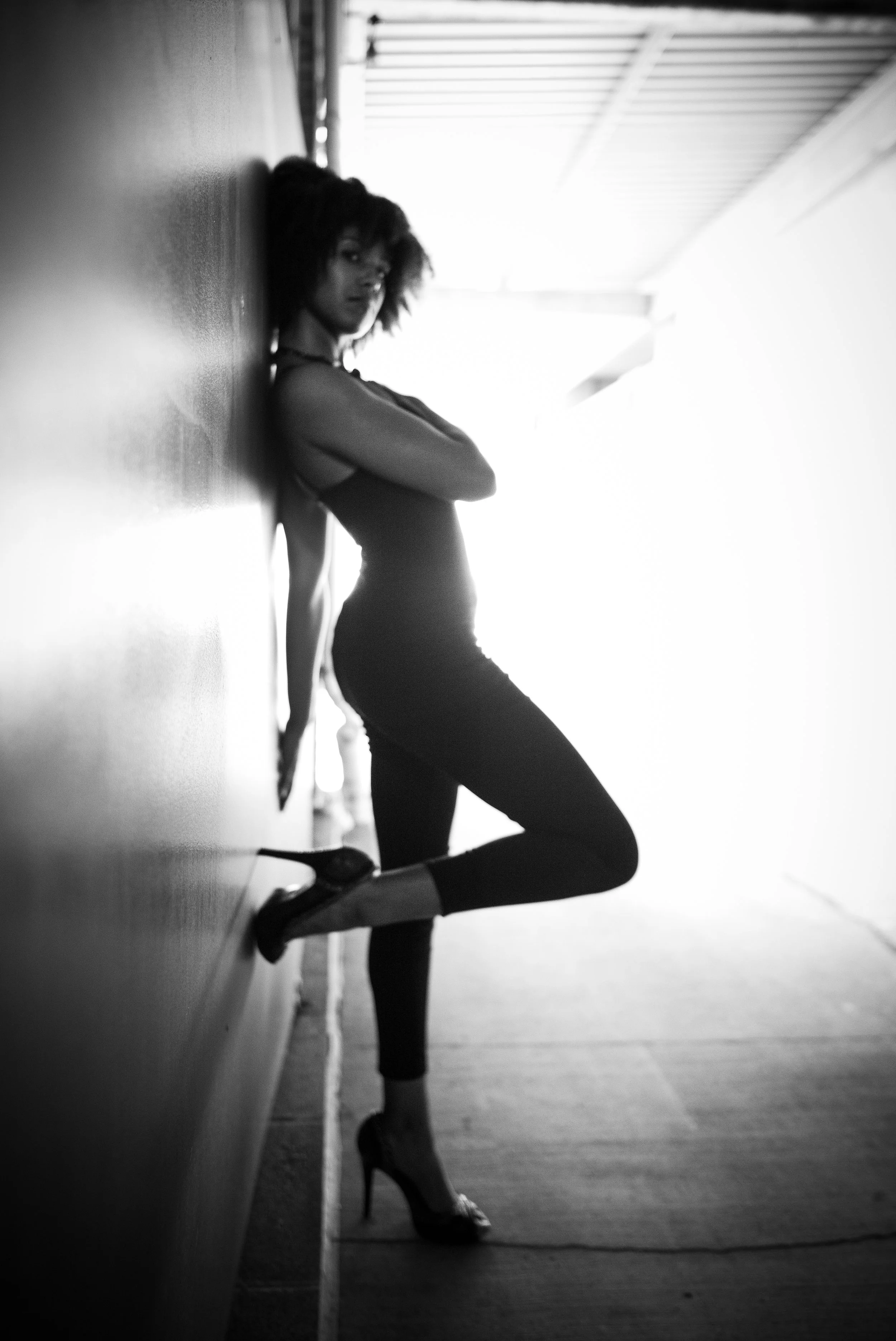 Black-and-white photo of a woman standing against a wall in a hallway, wearing high heels and a sleeveless top, with her arms crossed and one leg bent.