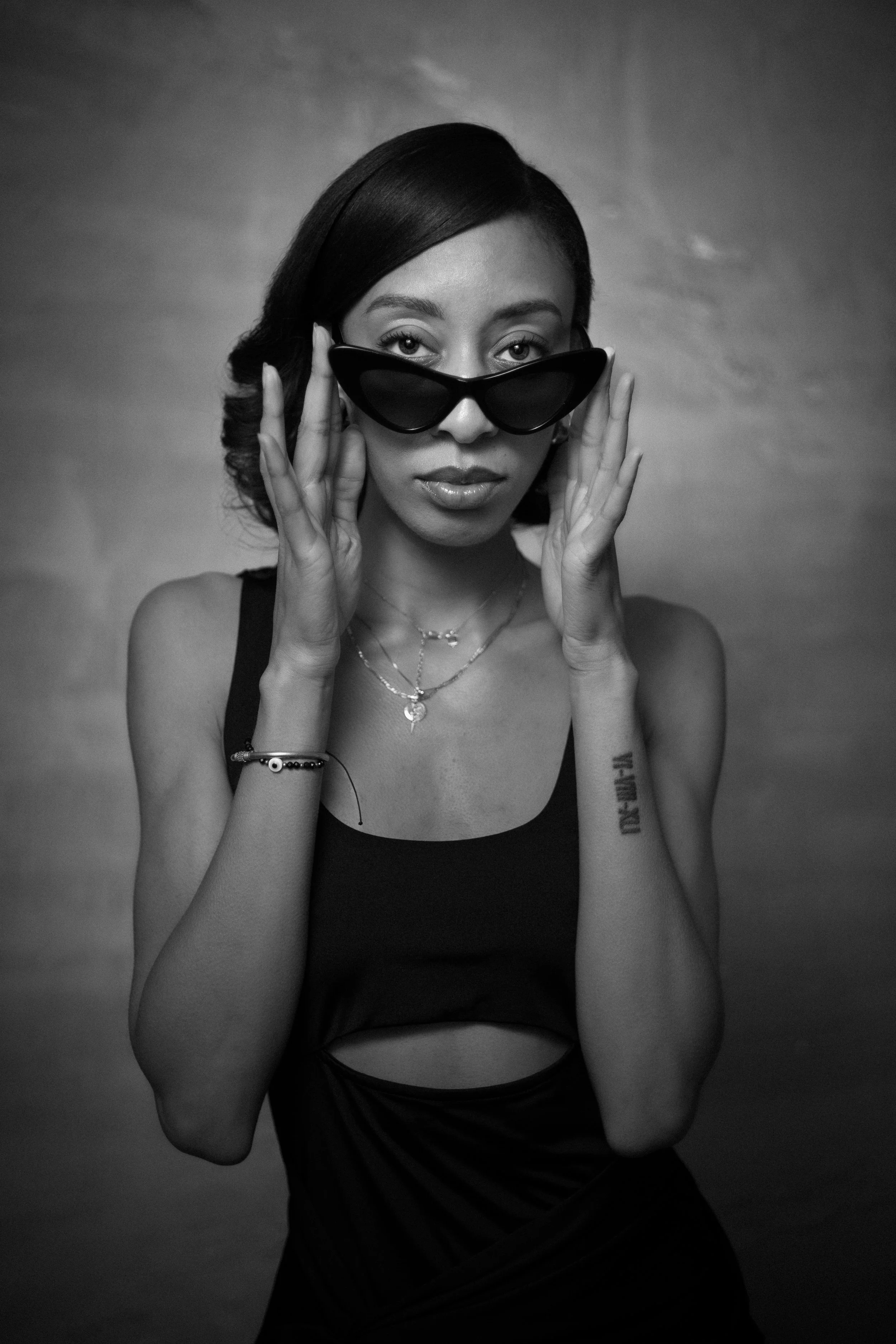 A woman with dark, wavy hair, wearing sunglasses, a sleeveless top, layered necklaces, and bracelets, posing with hands near her face against a plain background.