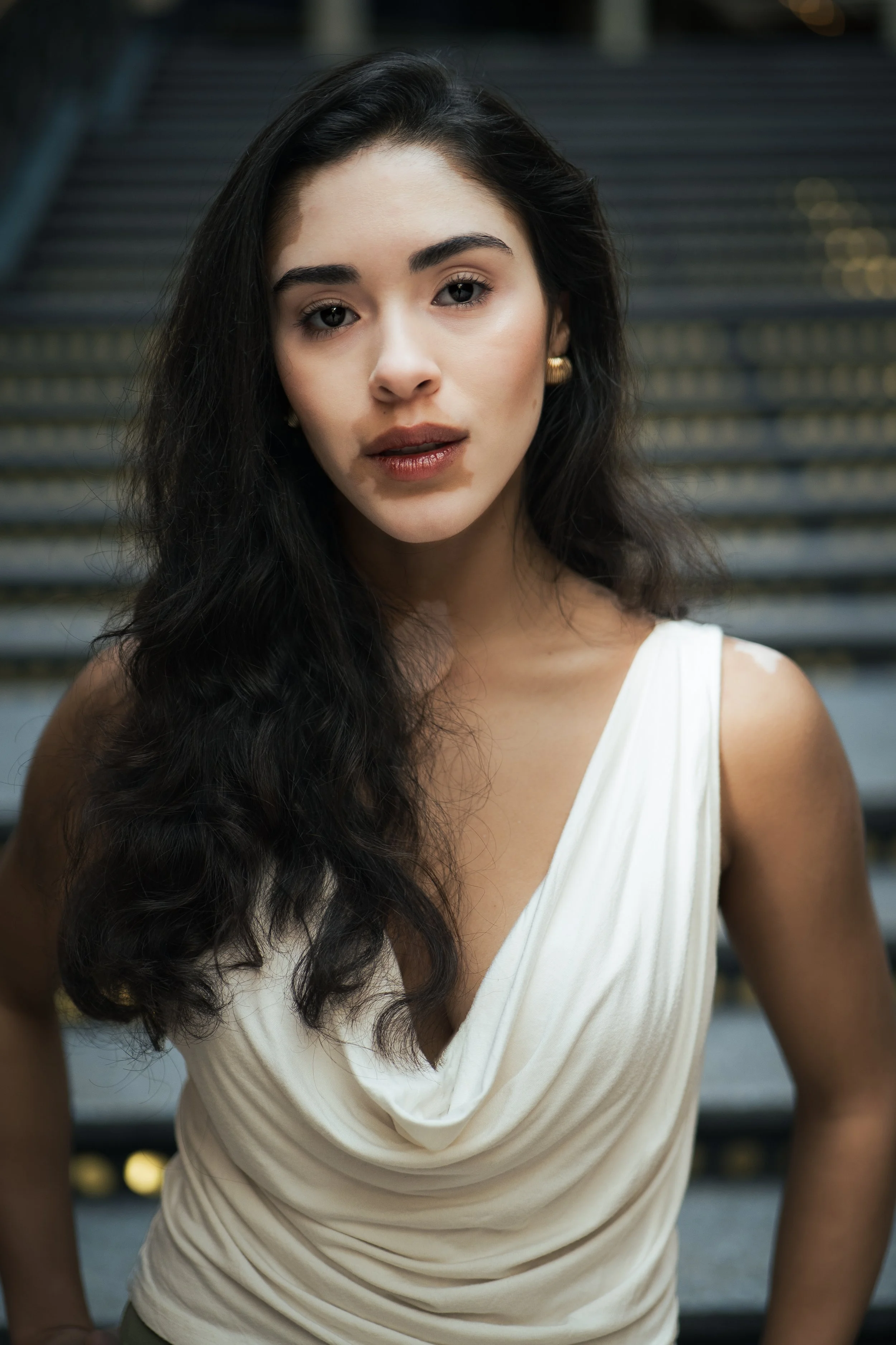 A young woman with long, wavy dark hair and light skin, wearing a sleeveless white top with draped fabric, standing in front of a staircase outdoors.