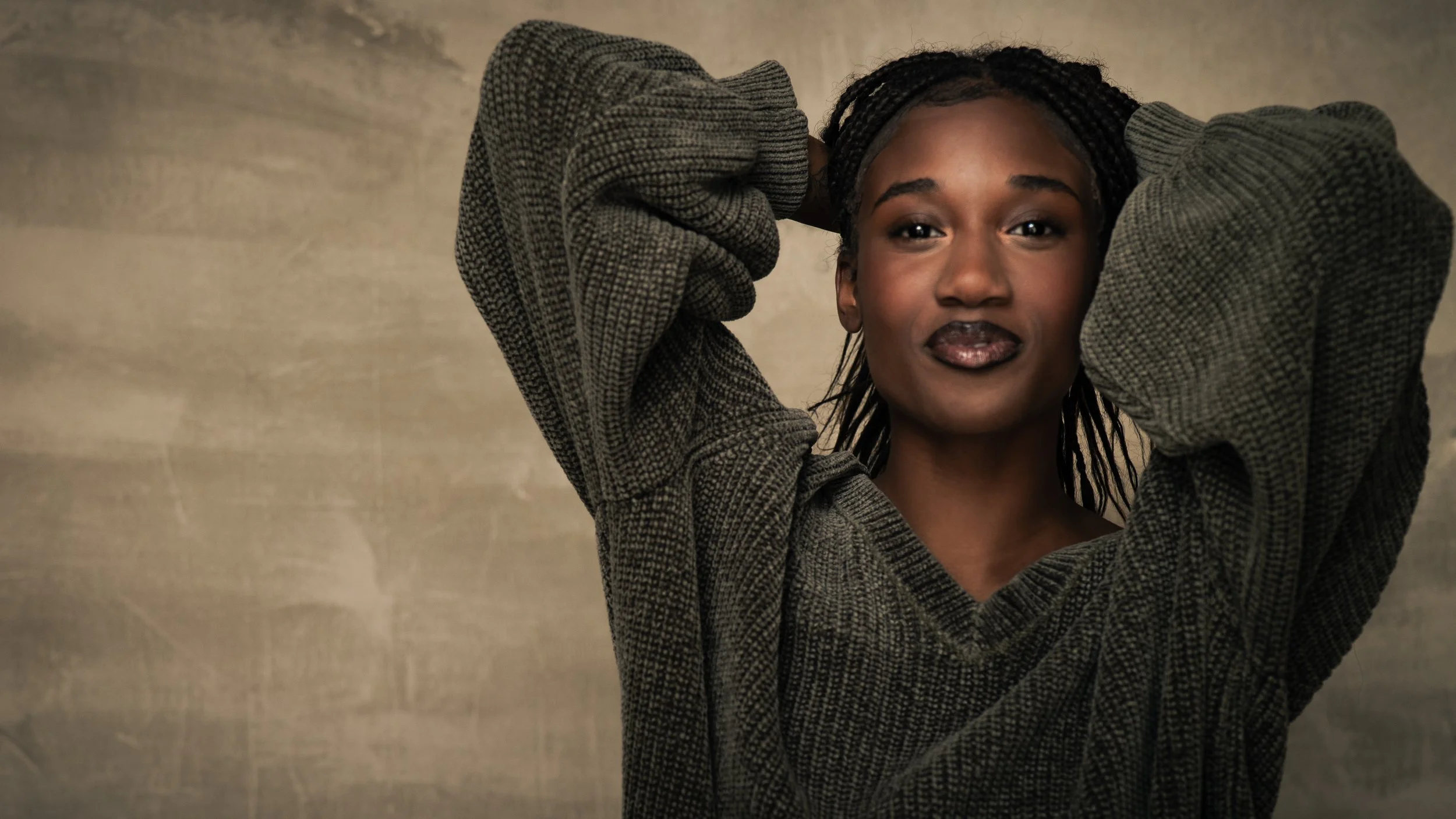 A woman with dark hair styled in braids, wearing a loose, oversized gray sweater, standing against a neutral background, looking directly at the camera with a confident expression.