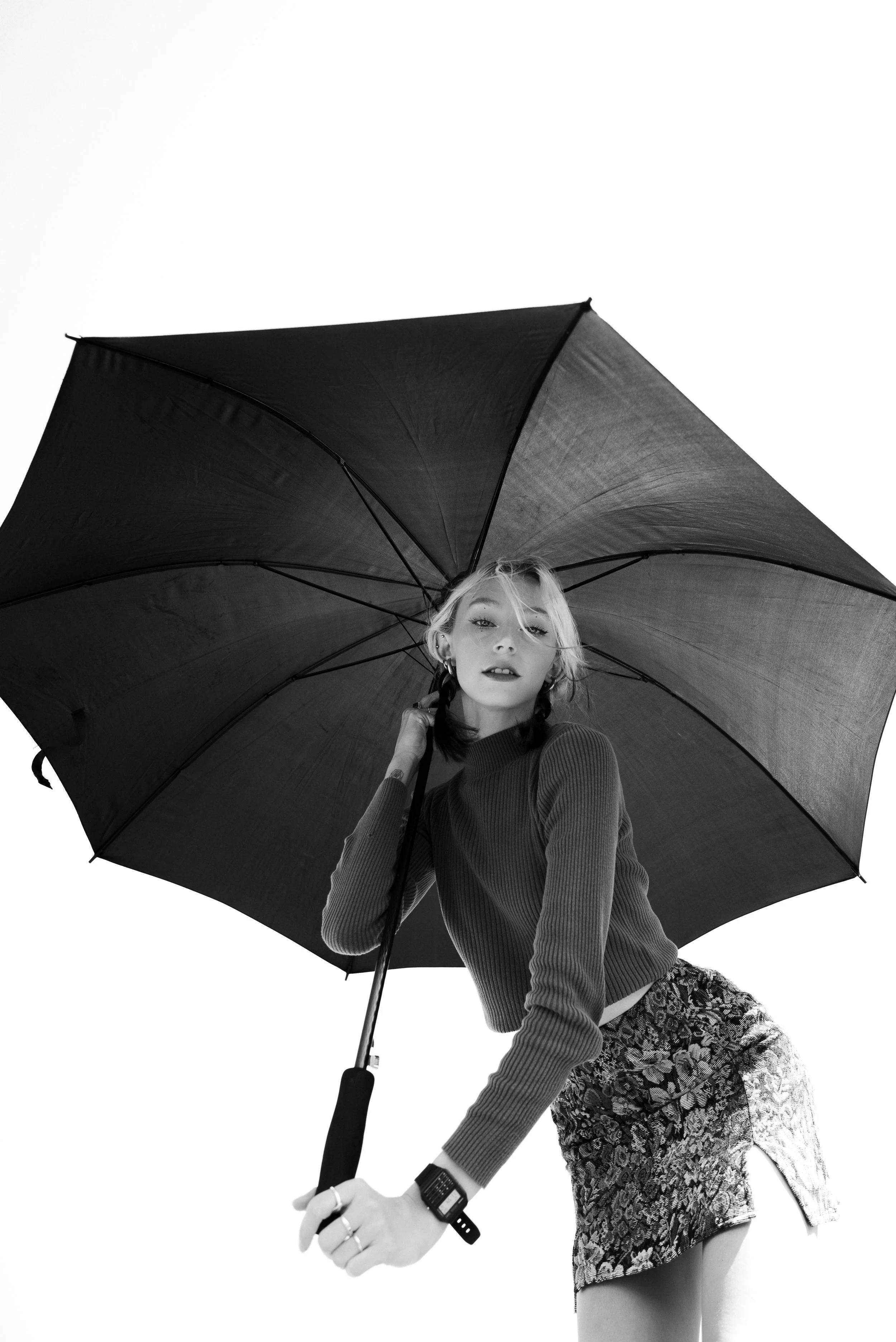 Black and white photo of a woman holding an umbrella, looking at the camera, wearing a long-sleeved top, a patterned skirt, and a watch.