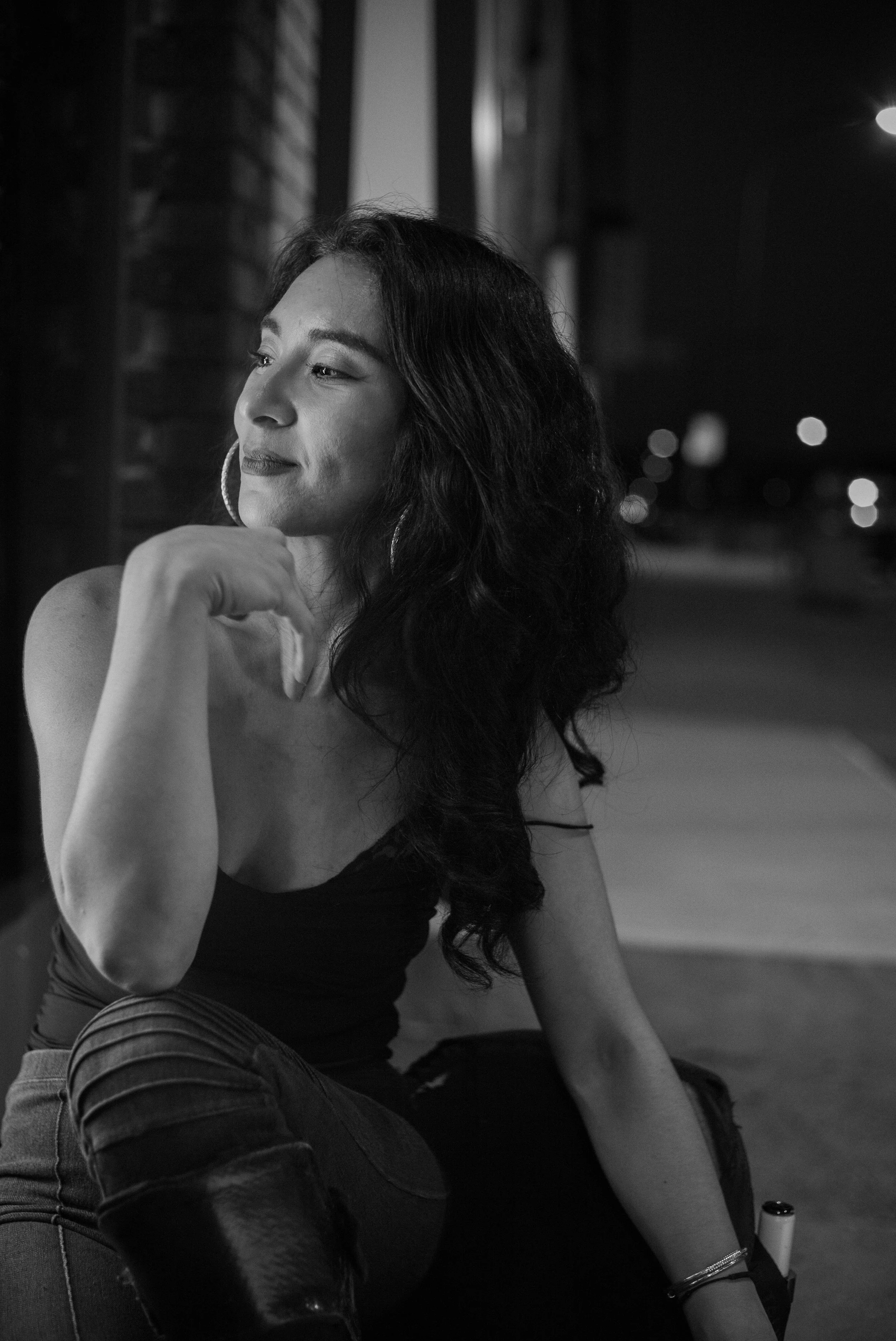 A woman with long, wavy hair sitting outside at night, wearing a sleeveless top and hoop earrings, gazing thoughtfully into the distance.