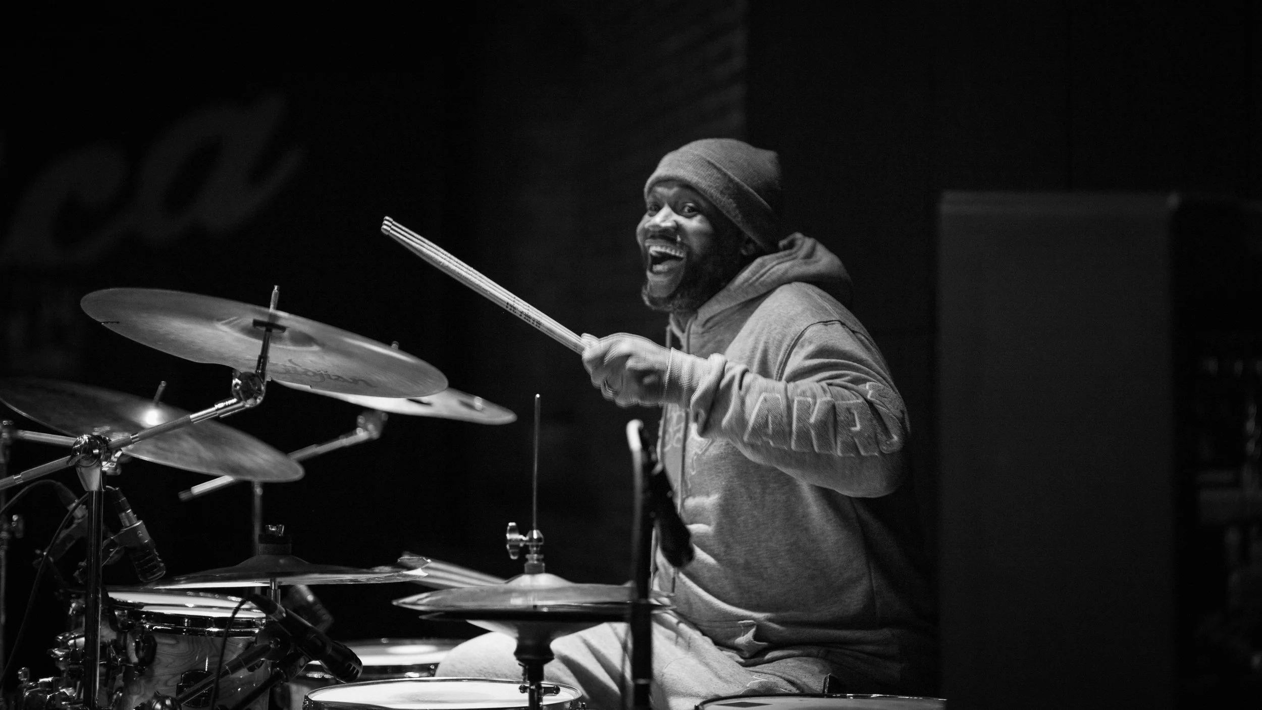 A man playing drums on stage, smiling and wearing a hoodie and beanie, in black and white.