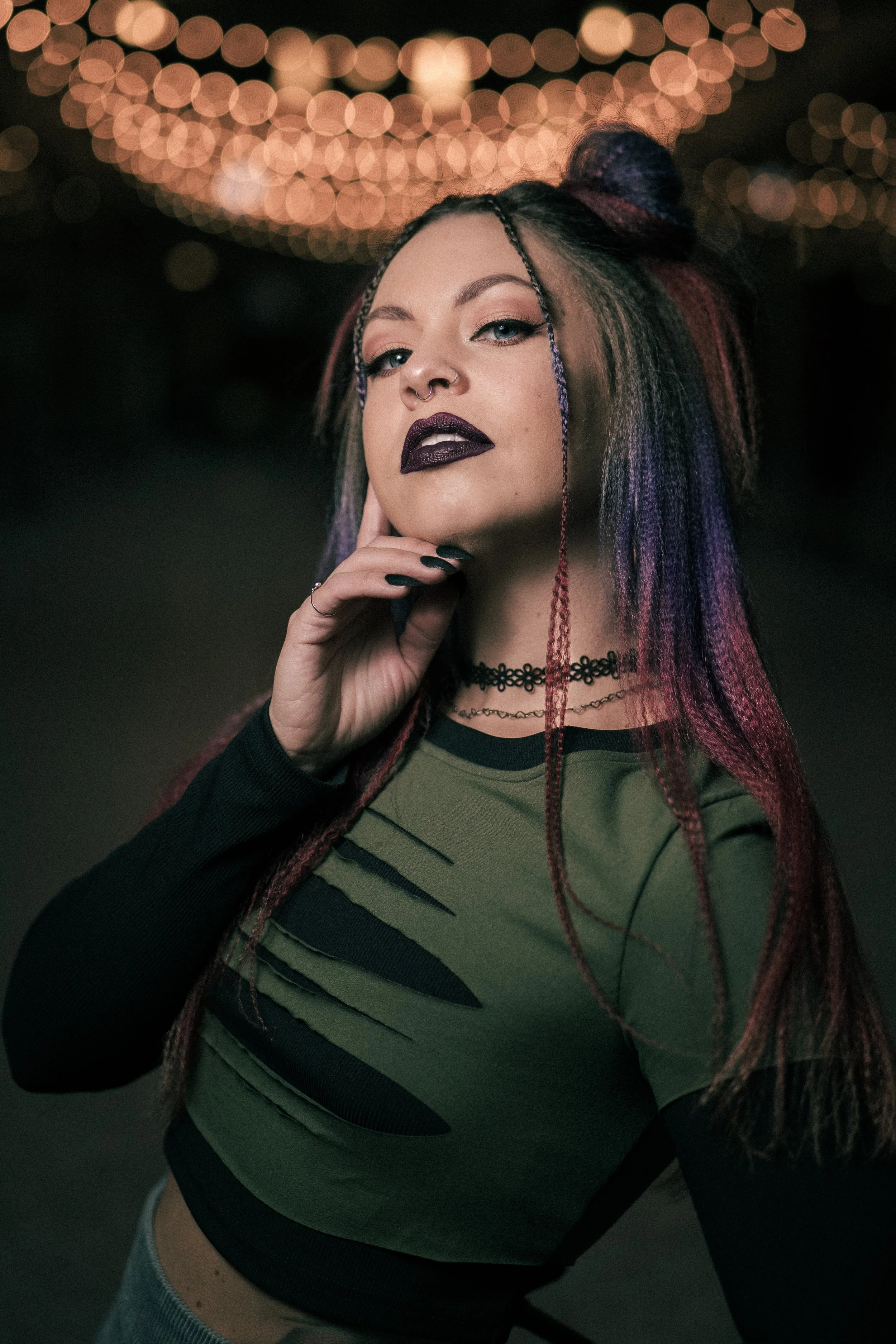 A young woman with colorful hair and dark makeup posing in front of a background with bokeh lights.