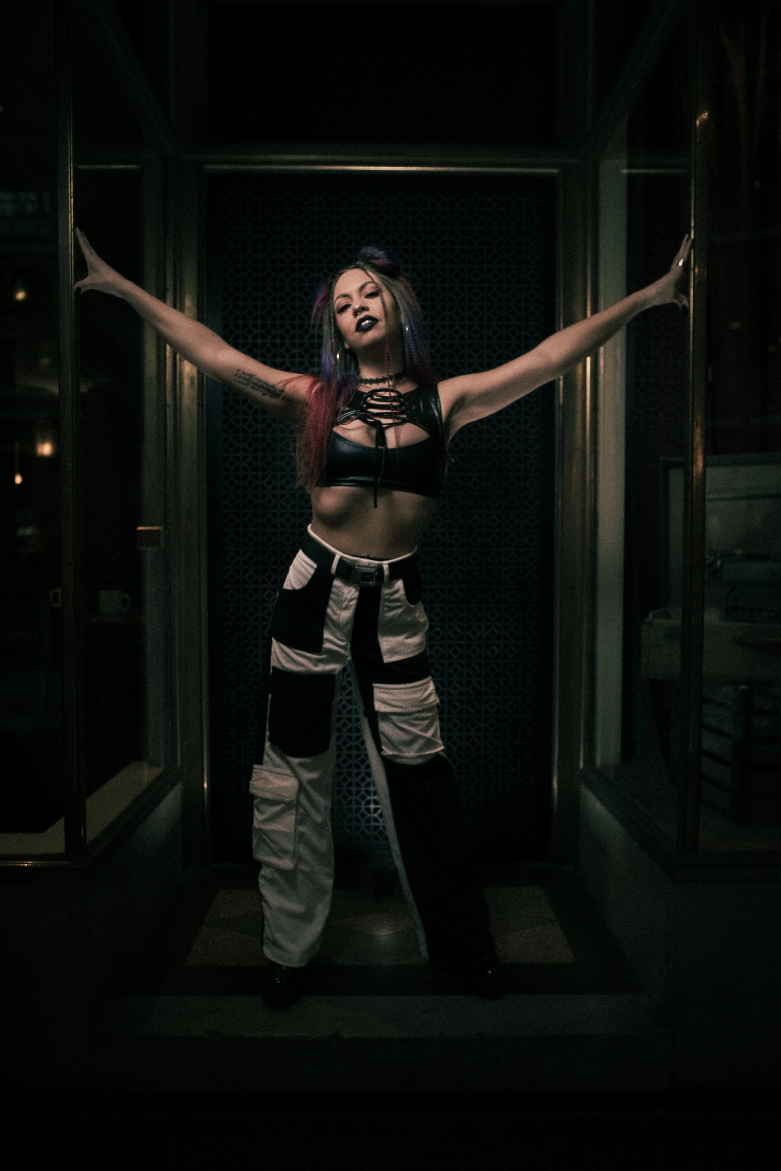 A woman with multi-colored hair and dark makeup posing with arms outstretched inside an elevator.