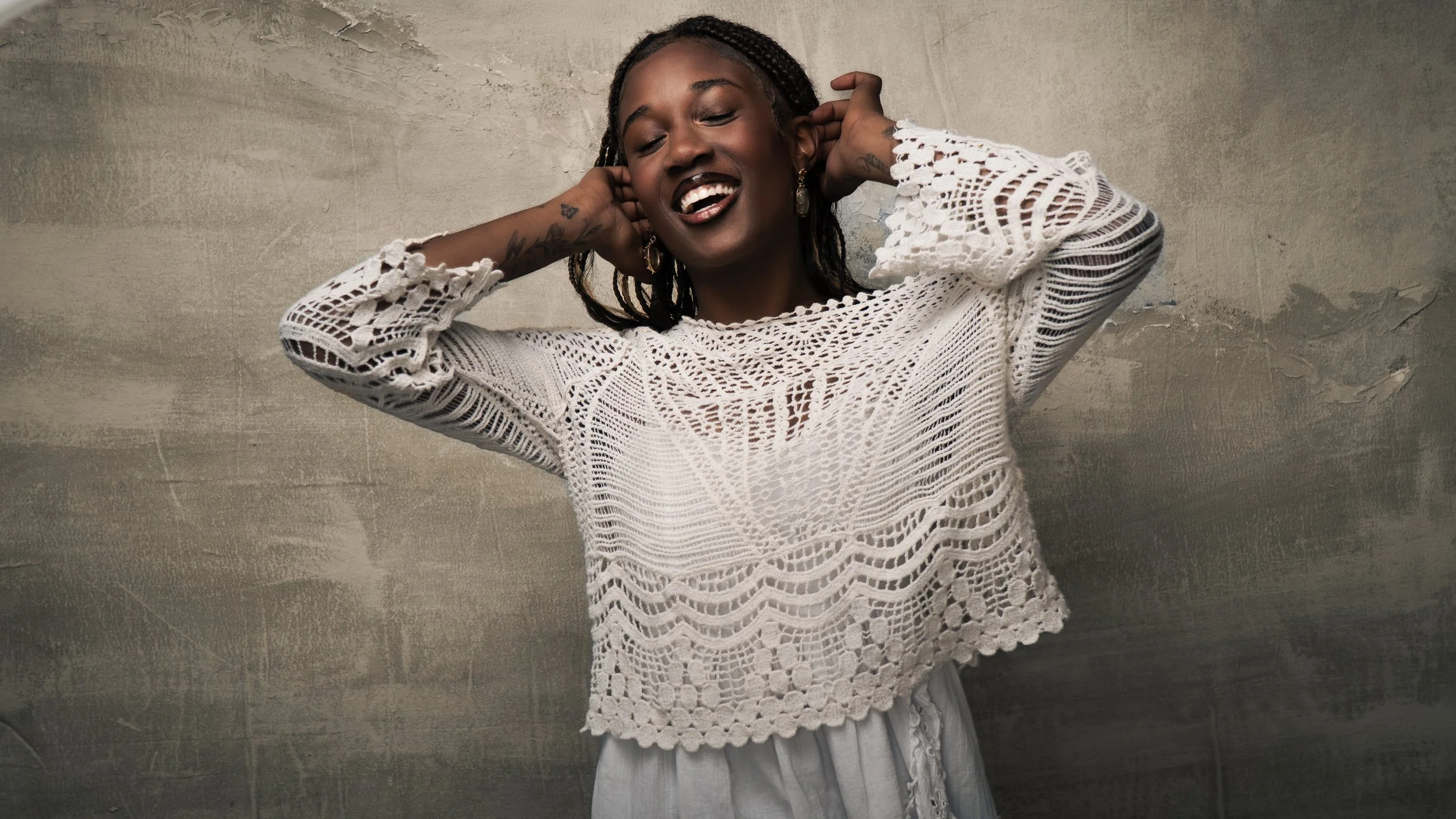 A smiling woman with dark skin and braids, wearing a white crocheted top and light-colored pants, stands against a textured gray wall, with her hands behind her ears.