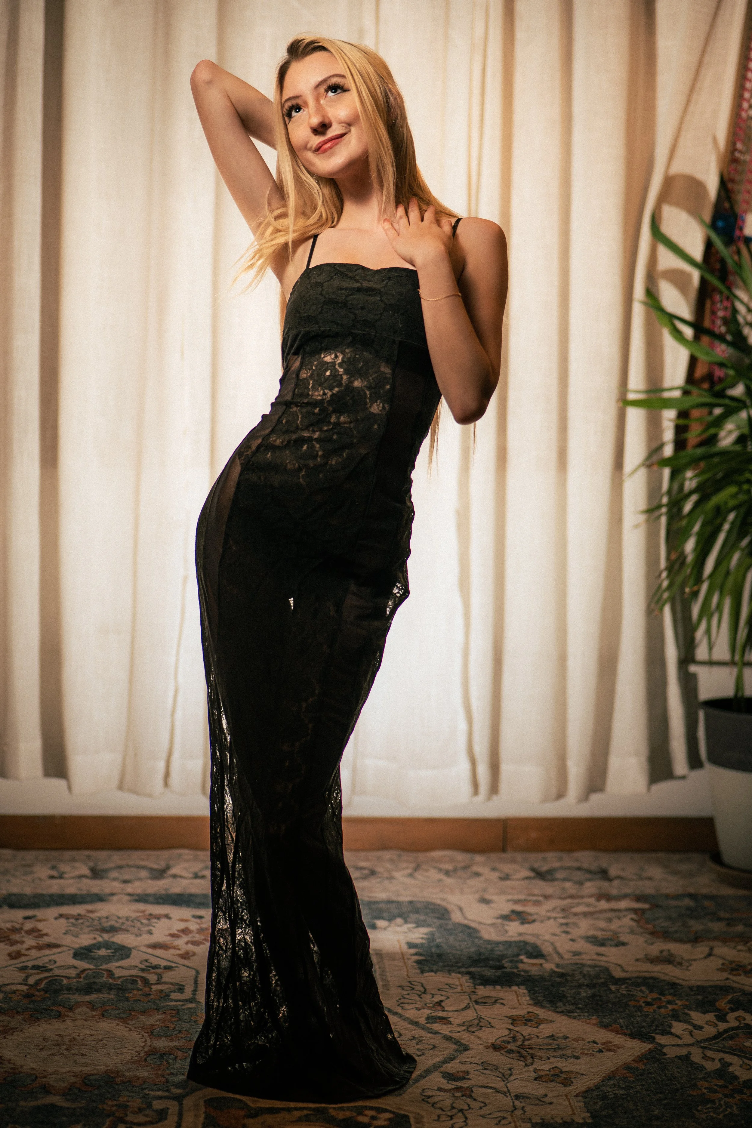 A woman in a black lace dress posing indoors in front of beige curtains with a potted plant nearby.