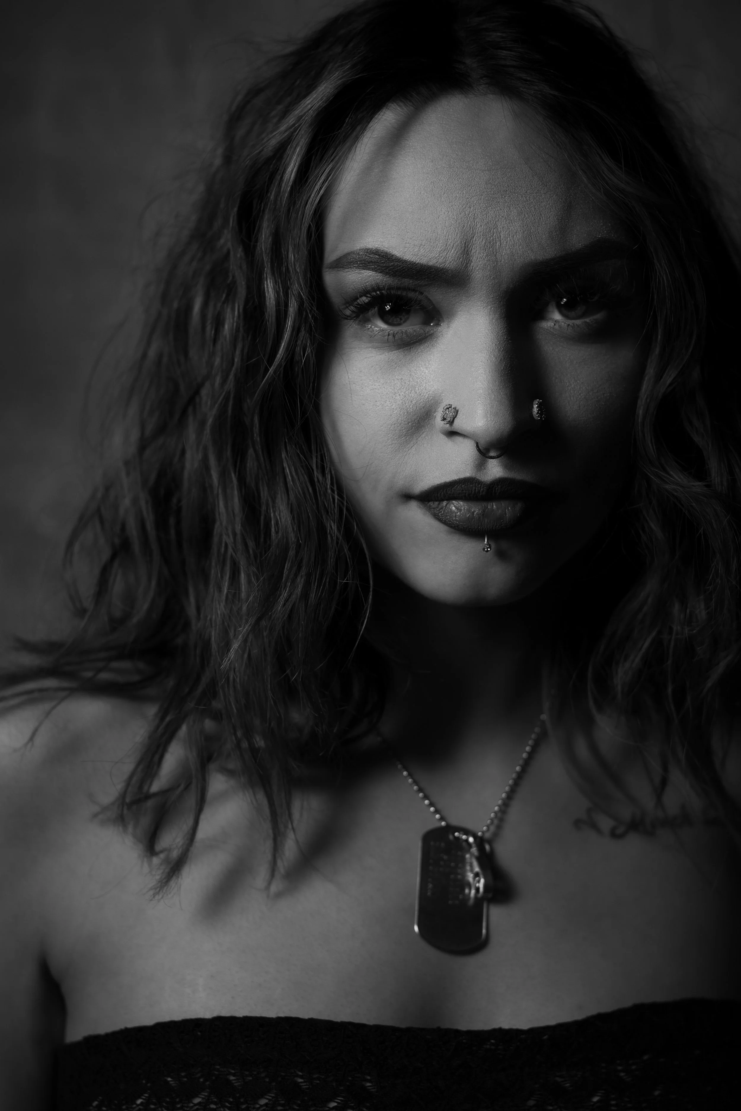 Black and white portrait of a woman with dark wavy hair, multiple facial piercings, and a serious expression, wearing a lace top and a dog tag necklace.