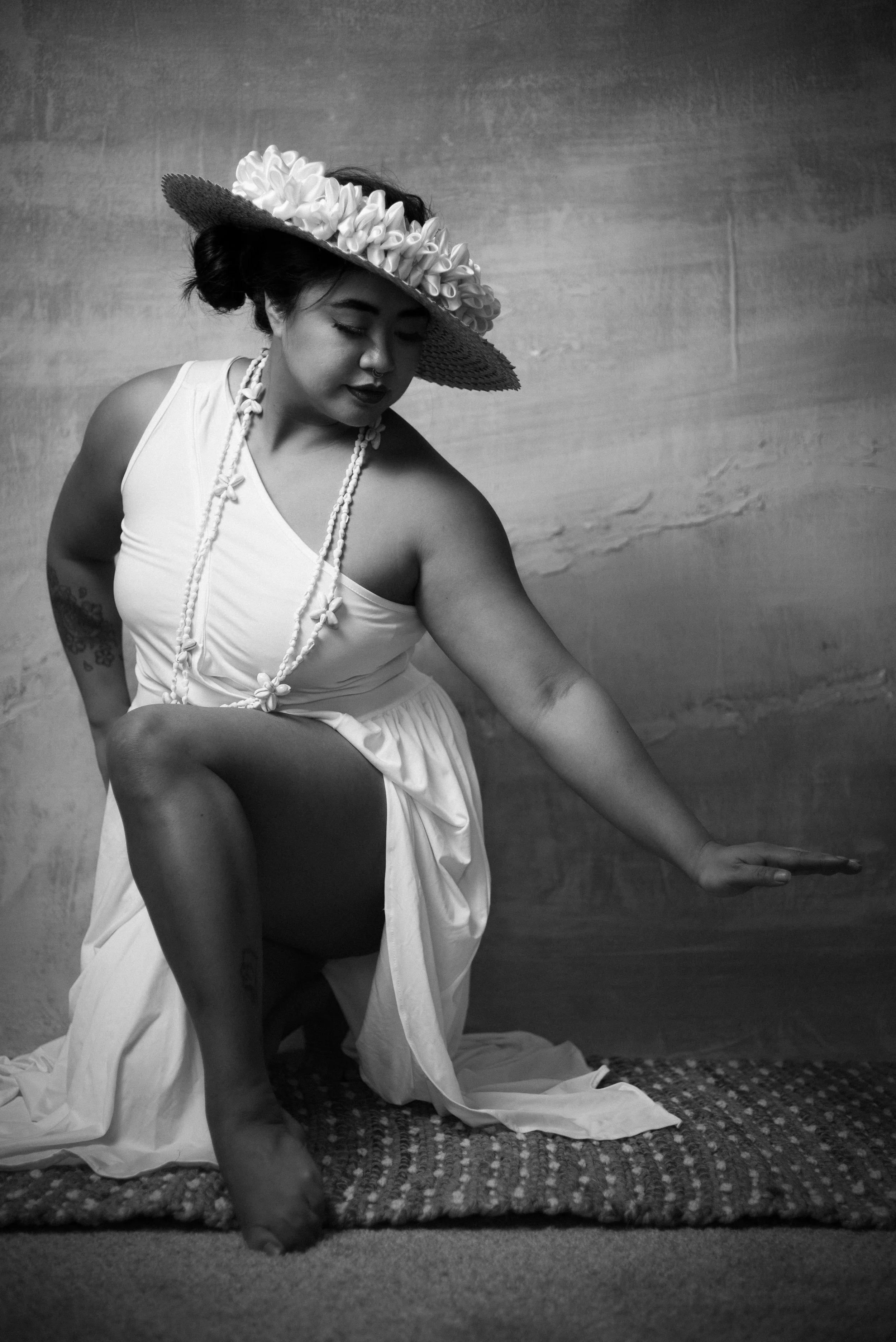 A woman in a white dress and large wide-brimmed hat with flowers poses with one knee raised, seemingly in a dance or artistic pose, on a textured rug against a plain background.