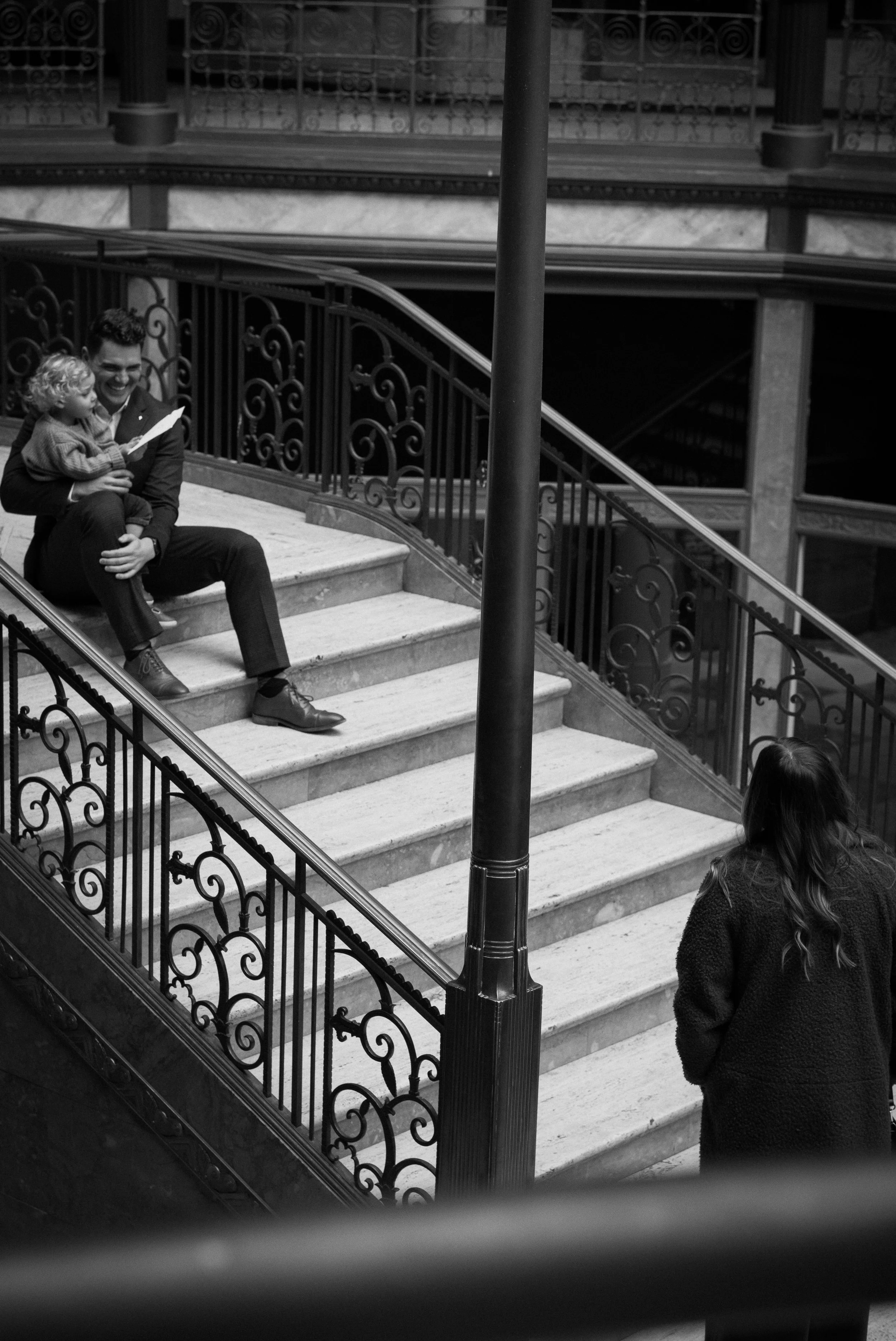A man sitting on a staircase holding a young child, both smiling and looking at a piece of paper, while a woman with long hair stands nearby, looking at them in an indoor setting with ornate railings and marble steps.
