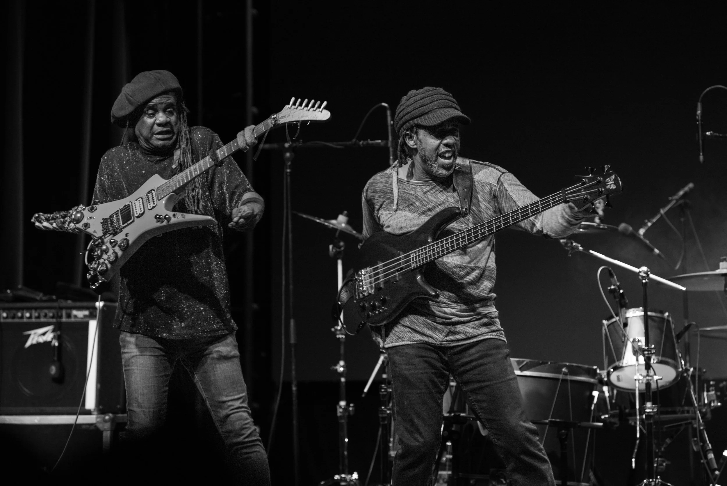 Black and white photo of two musicians performing on stage. One is playing an electric guitar and wearing a cap, while the other is playing a bass guitar and wearing a long-sleeve shirt and a cap. There are drums and microphones in the background.