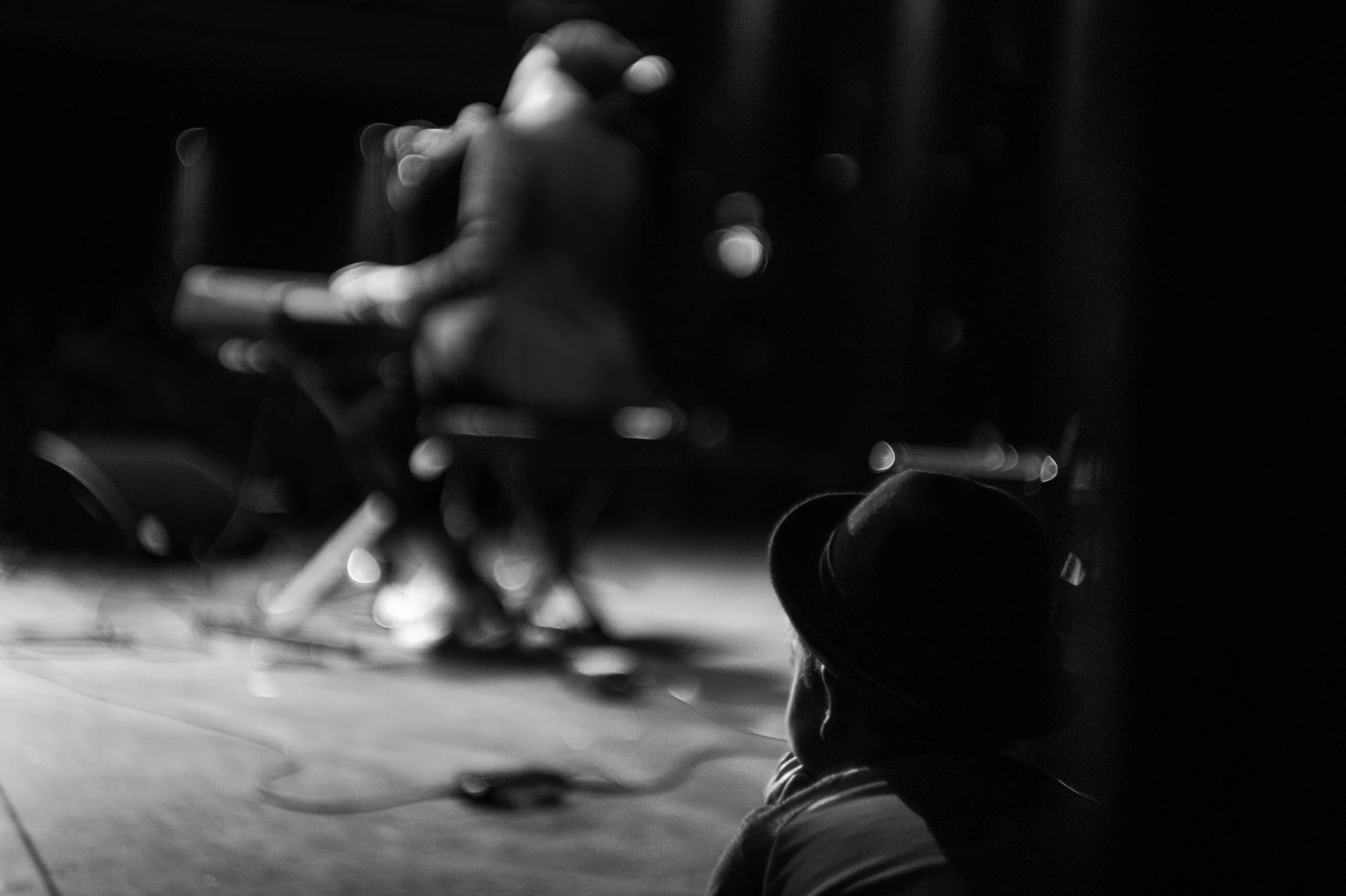 A person in a fedora hat sitting on the floor in a dark room, facing a blurred performer on stage, who is playing a guitar.