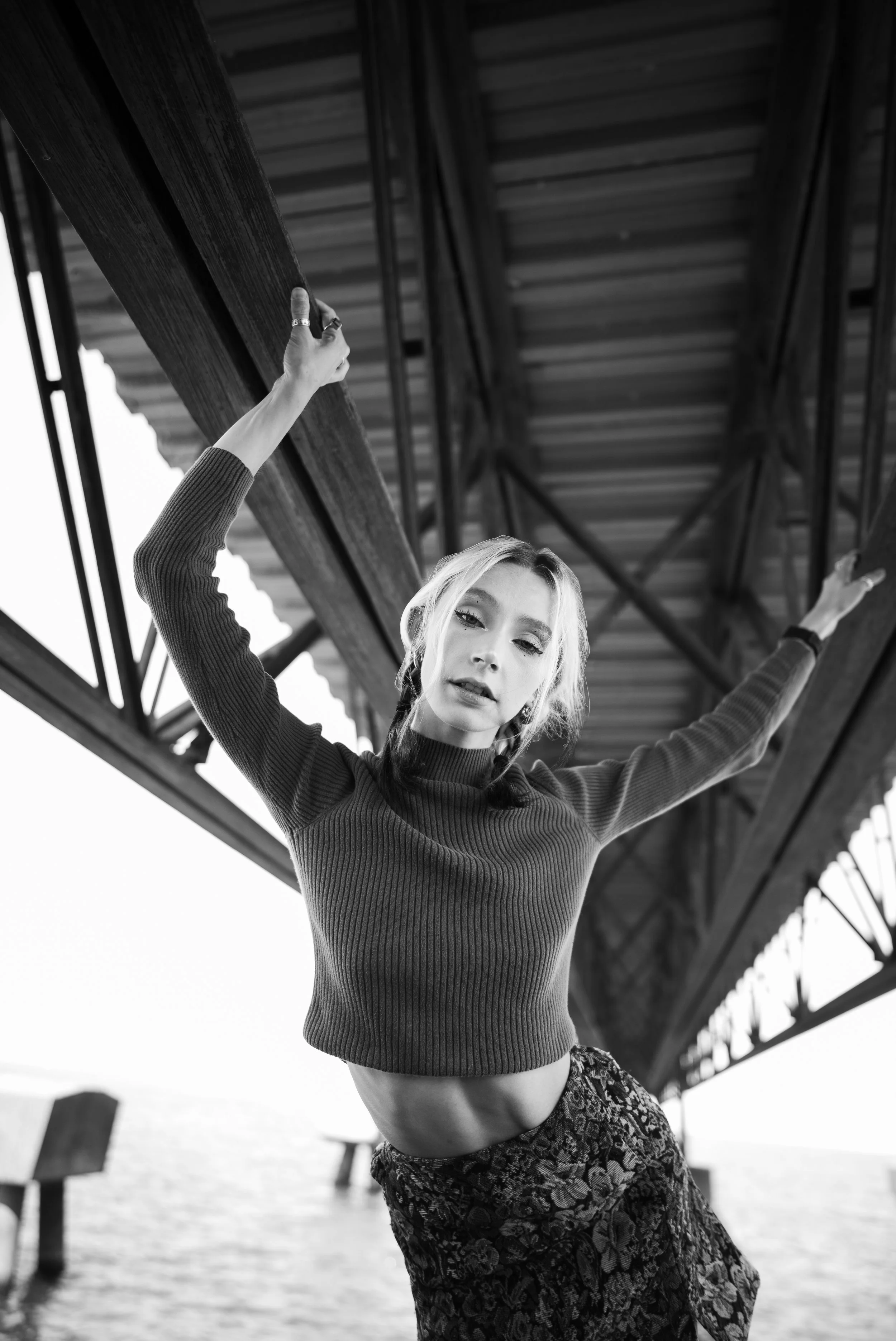 A woman standing under a bridge, holding onto the structure with both hands, with water and the bridge in the background.