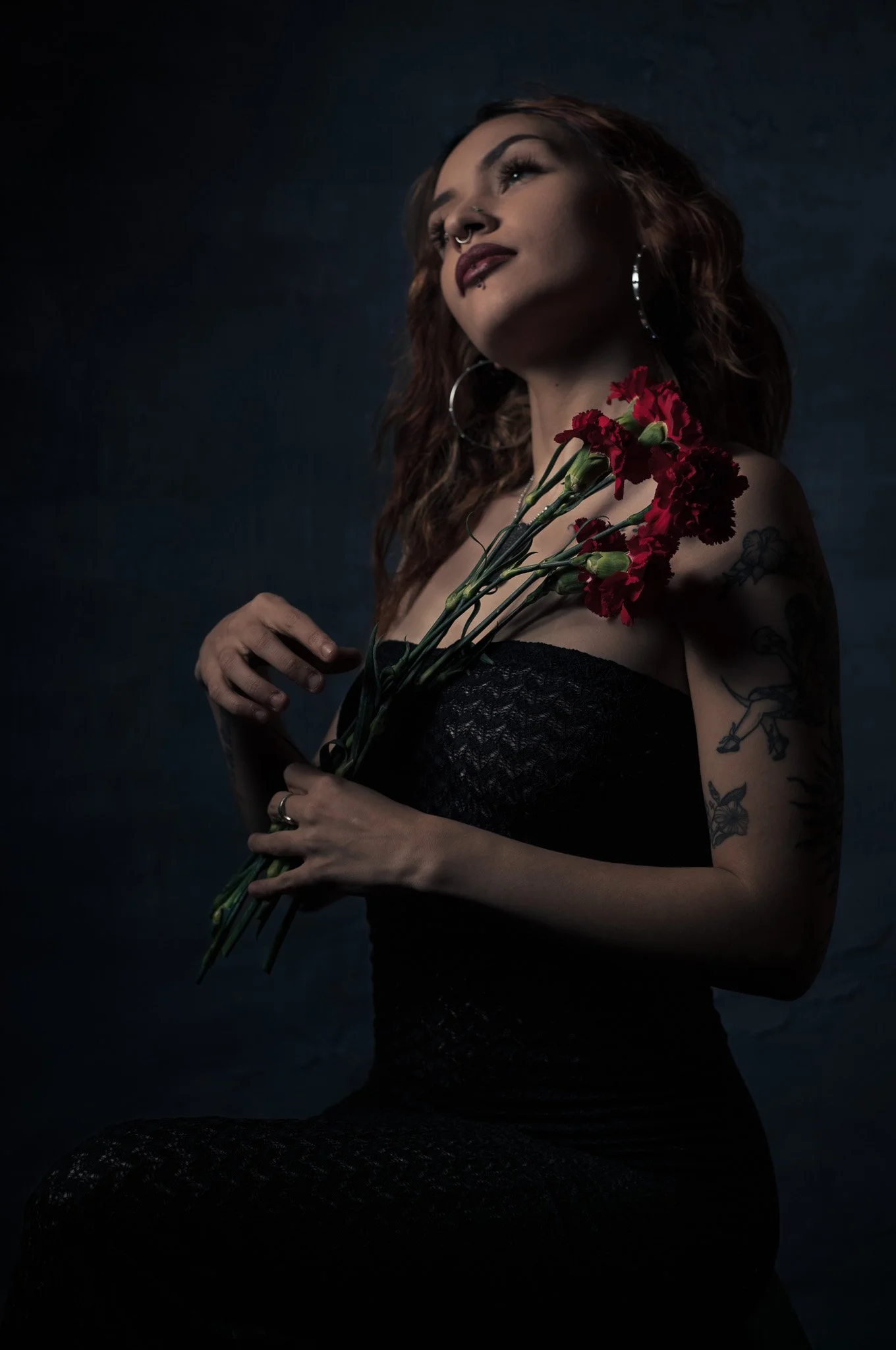 A woman with long, wavy hair and tattoos on her arm, wearing a strapless black dress, holding a bunch of red flowers against a dark background.