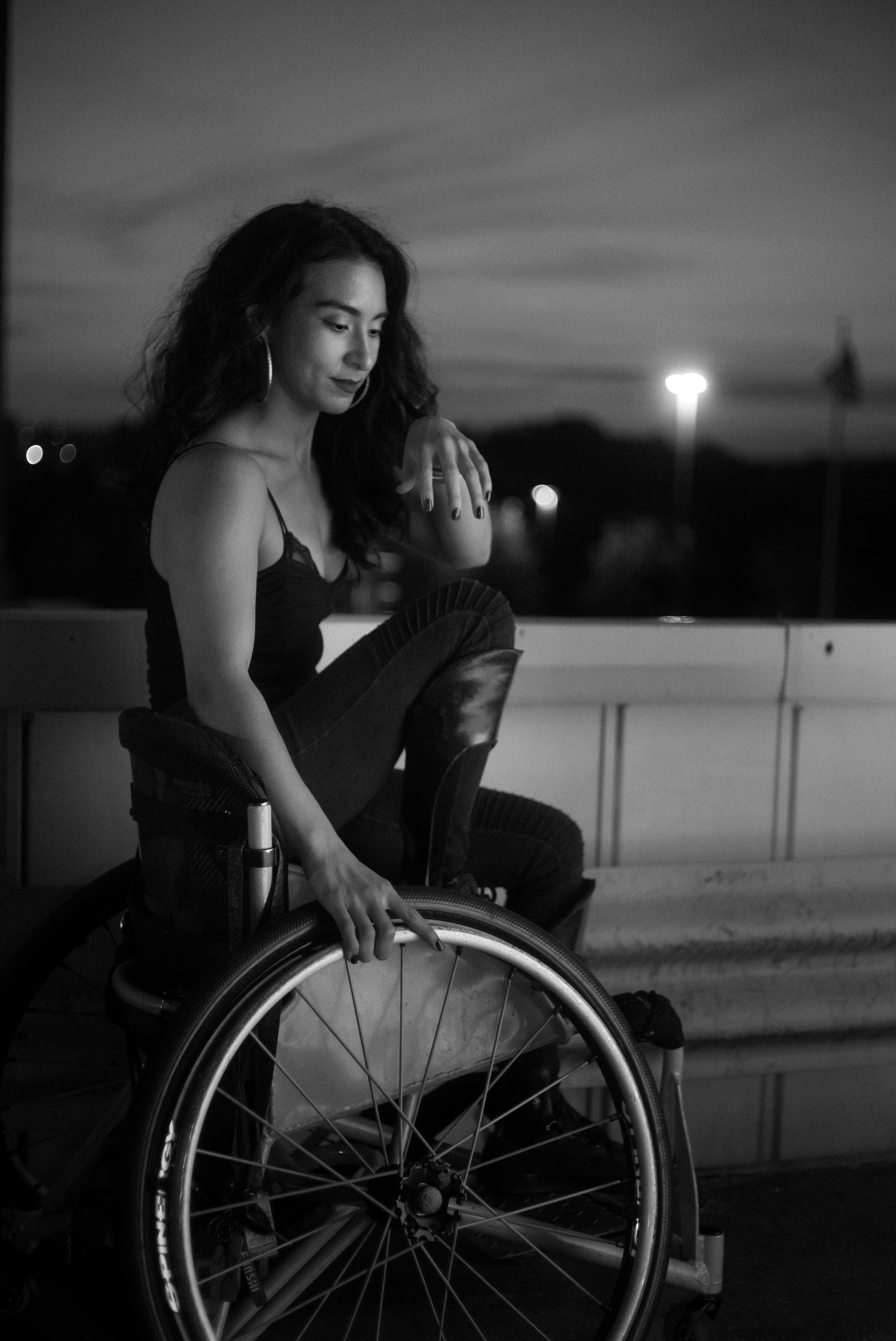 A woman with long wavy hair, wearing hoop earrings, a sleeveless top, and jeans, sitting in a wheelchair outdoors during evening or night, with her hand touching her knee. The background shows blurred lights and a flag, under a cloudy sky.
