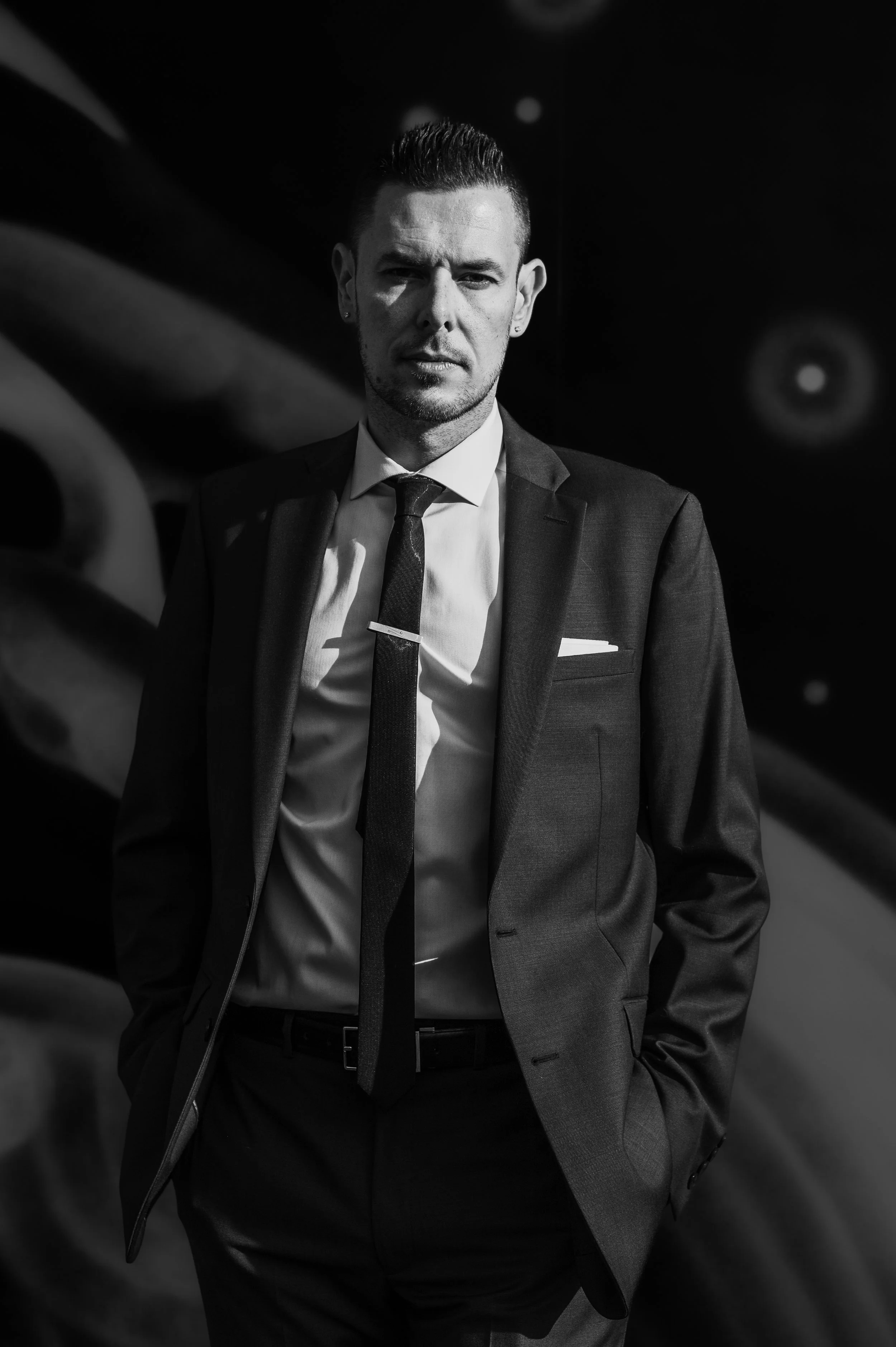 A man in a suit and tie standing with hands in pockets, looking serious, in black and white.