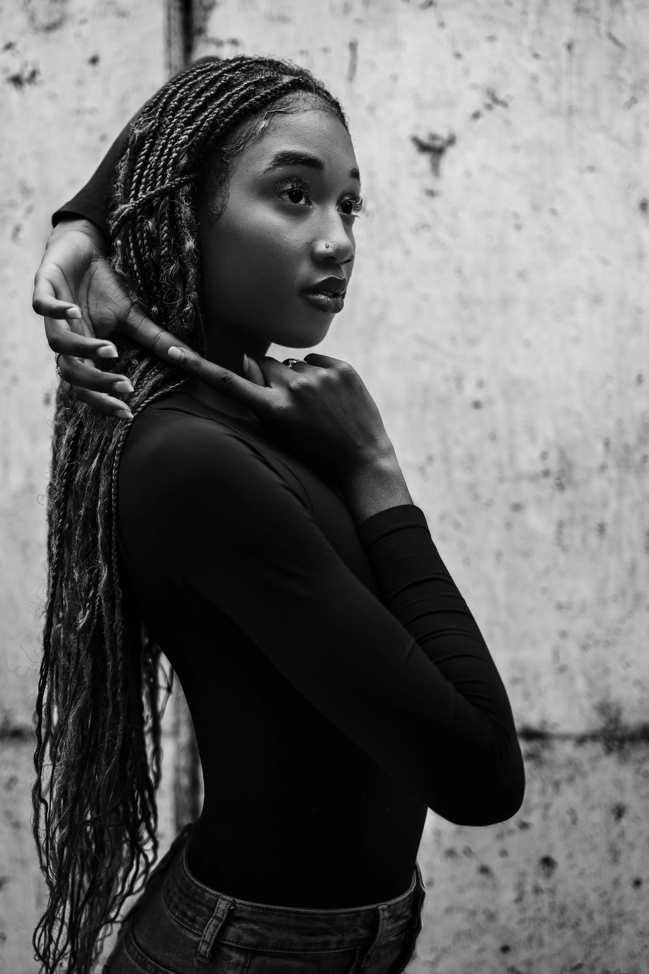 A young woman with long braided hair posing against a textured wall, looking thoughtfully into the distance.