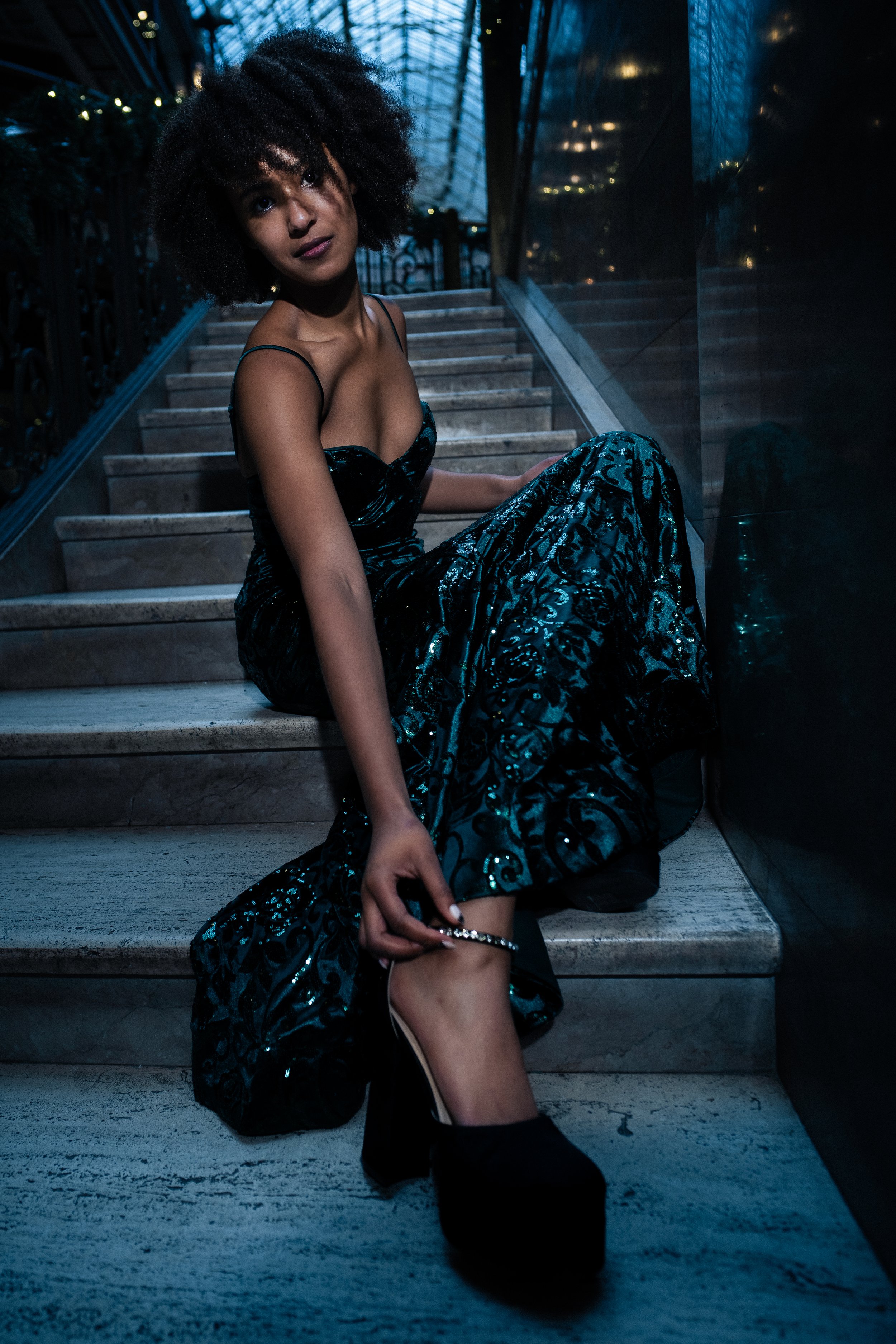 A woman in a dark, shiny, patterned evening gown sits on a staircase, adjusting her black high-heeled shoe with a jewelry anklet in a dimly lit setting.