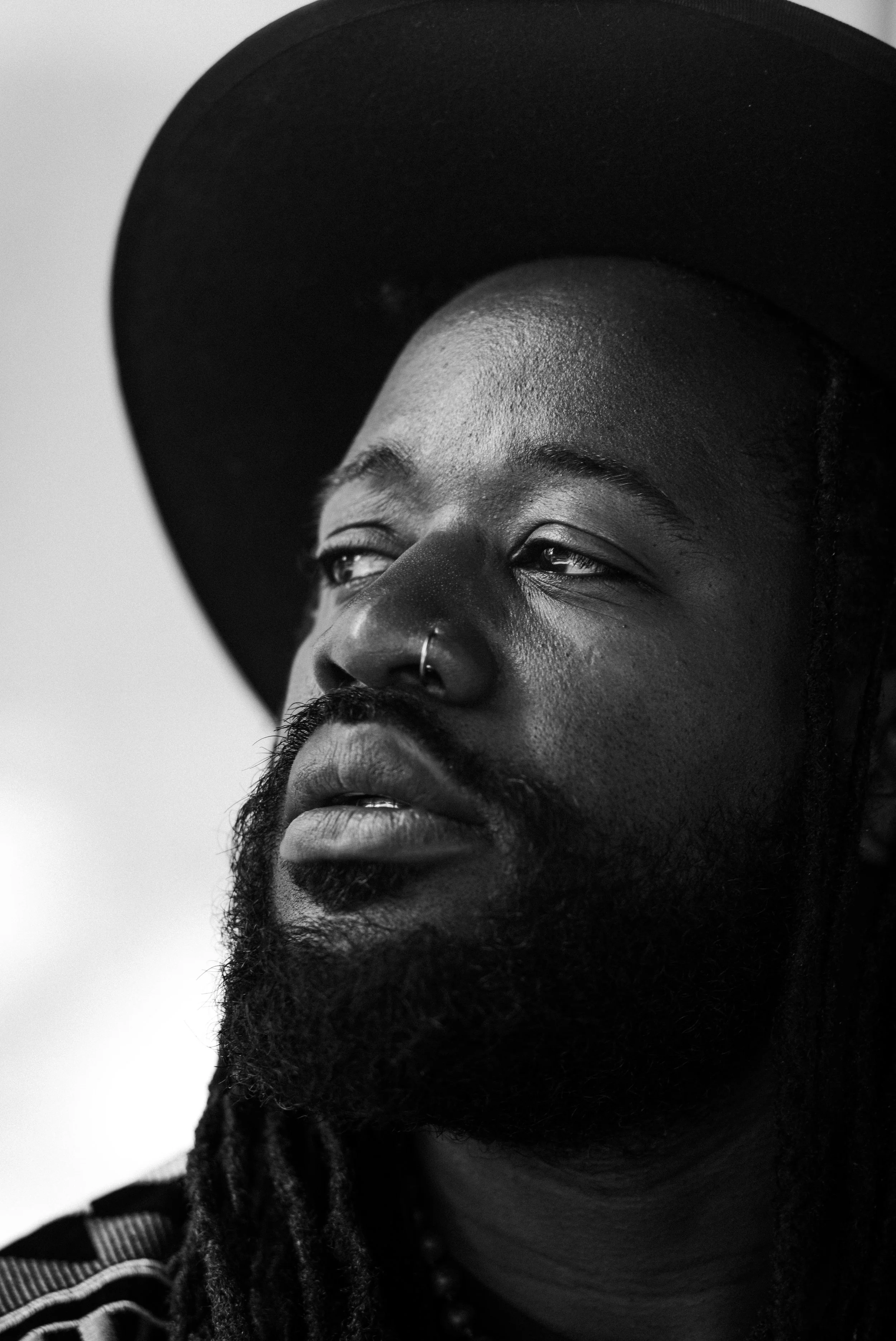 Close-up black and white portrait of a man with a beard, wearing a wide-brimmed hat, with his eyes partly closed and a nose ring.