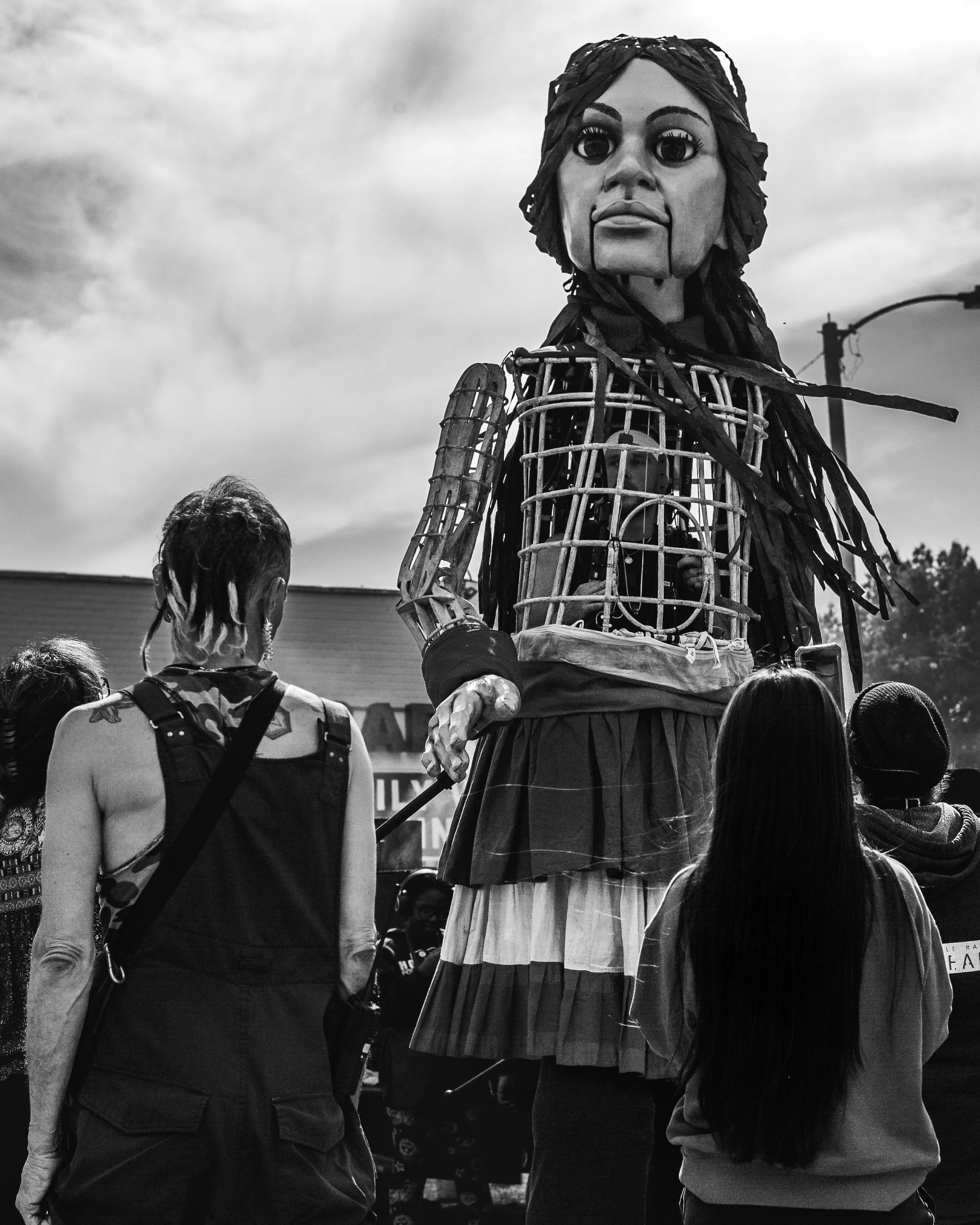 Crowd of people gathered around a large puppet of a woman with a big head, long hair, and expressive eyes, made of wood and fabric, during a daytime outdoor event.