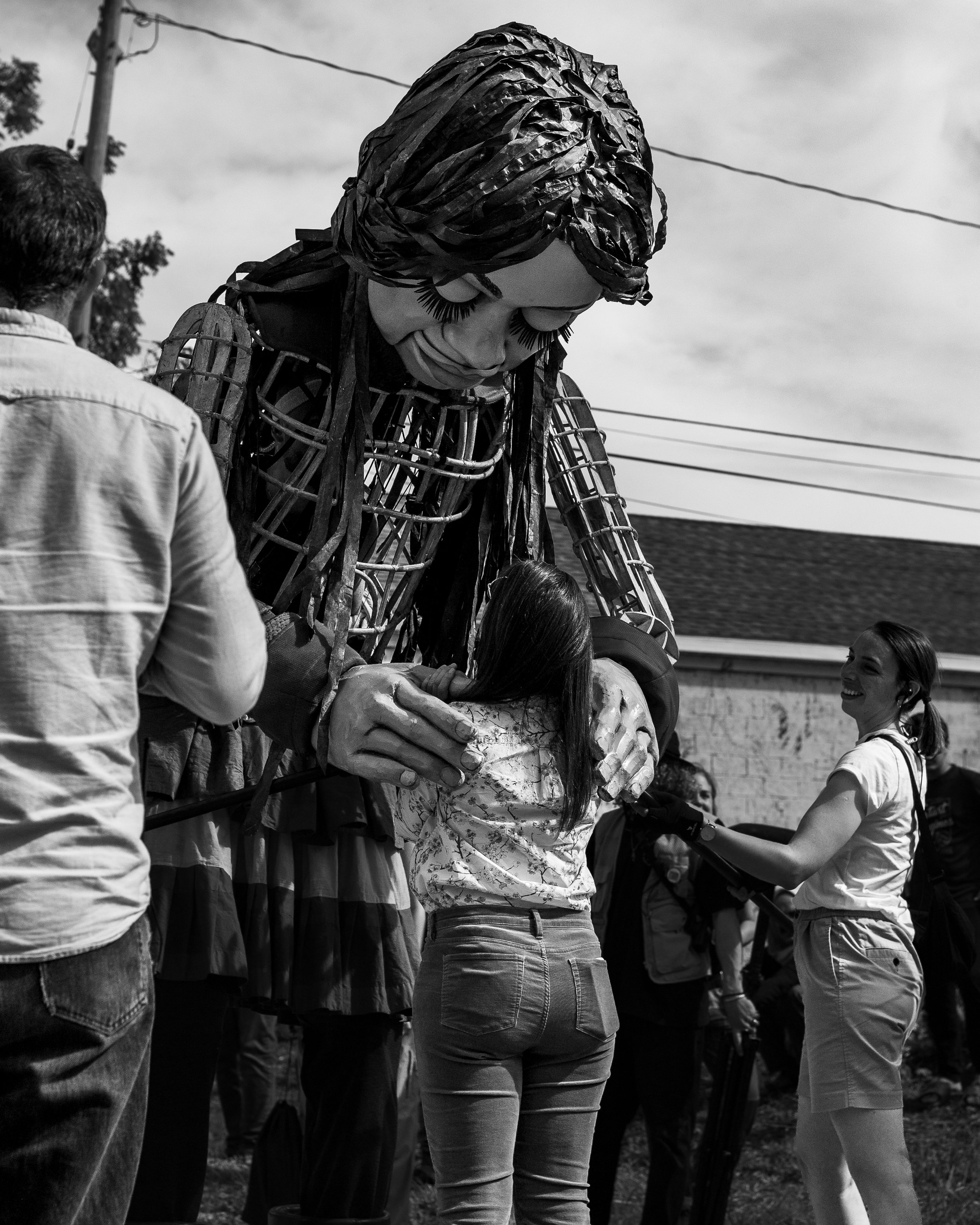 A large puppet of a woman with long hair, made of metal and other materials, bows down to hug a young girl.