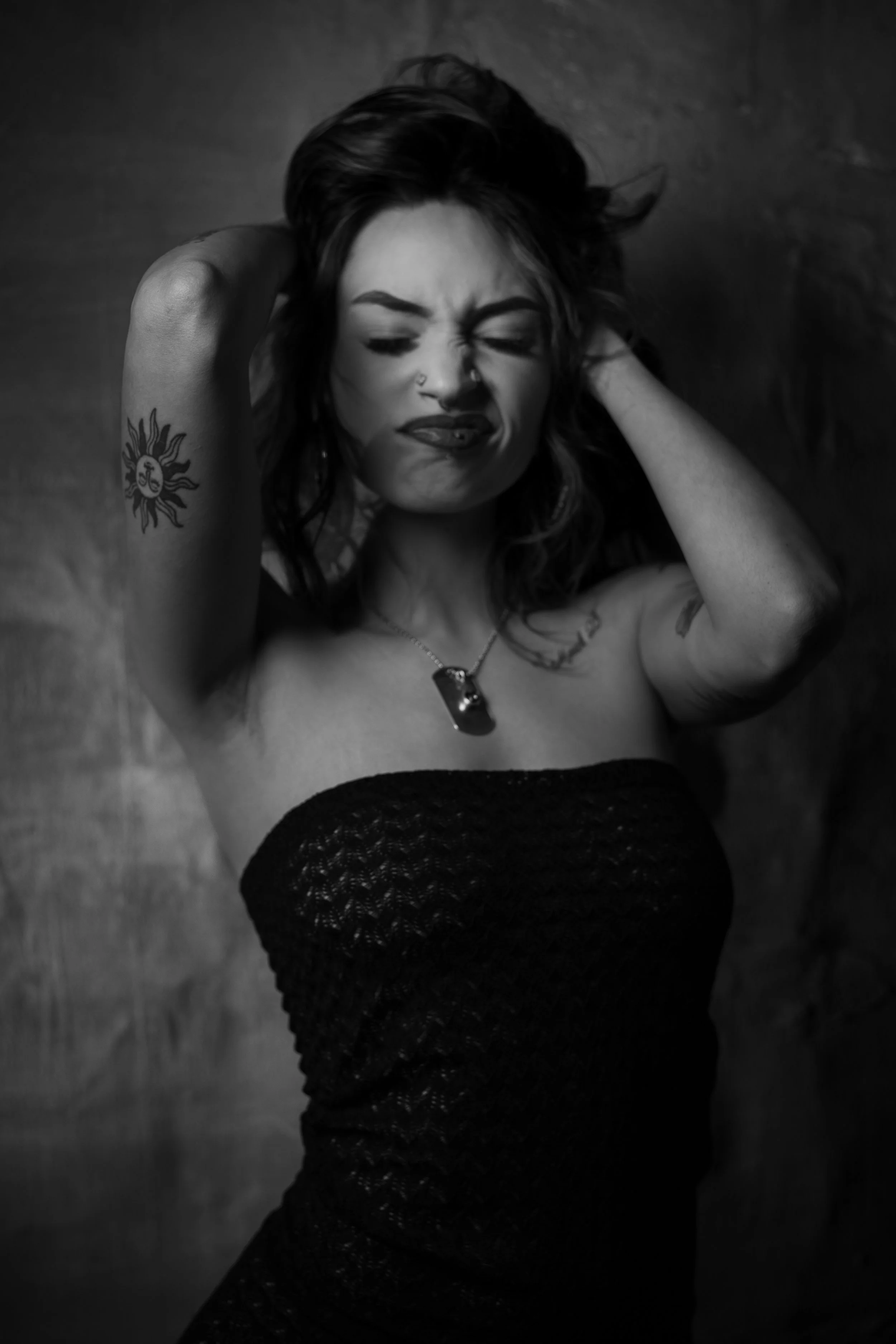 Black and white photograph of a woman with dark, wavy hair, tattoos, a nose piercing, and a gemstone necklace, wearing a strapless textured dress, with her eyes closed and her hands behind her head.