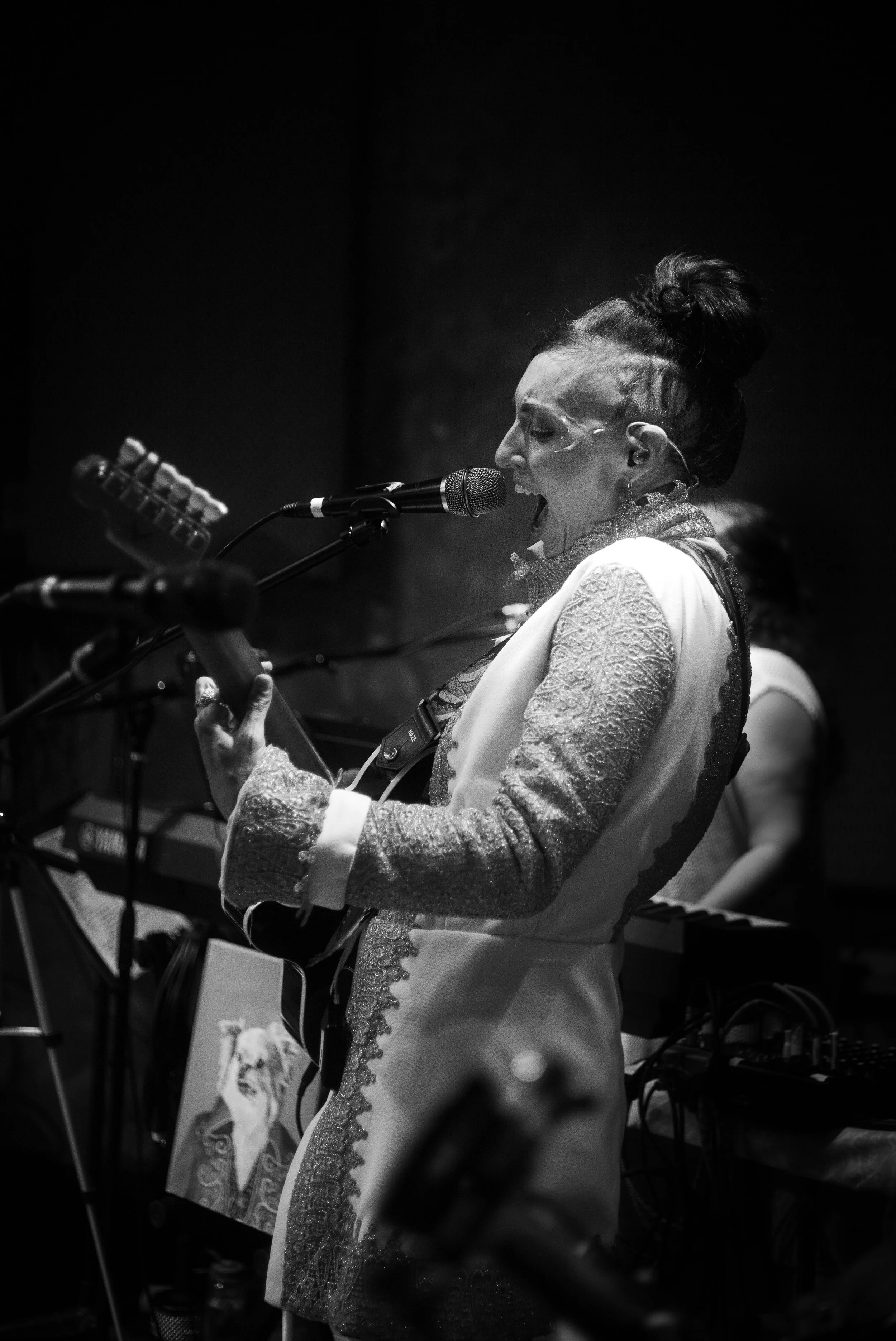 A woman singing passionately into a microphone while playing an acoustic guitar on stage. She has her hair styled in an updo and is wearing a textured jacket with lace details.