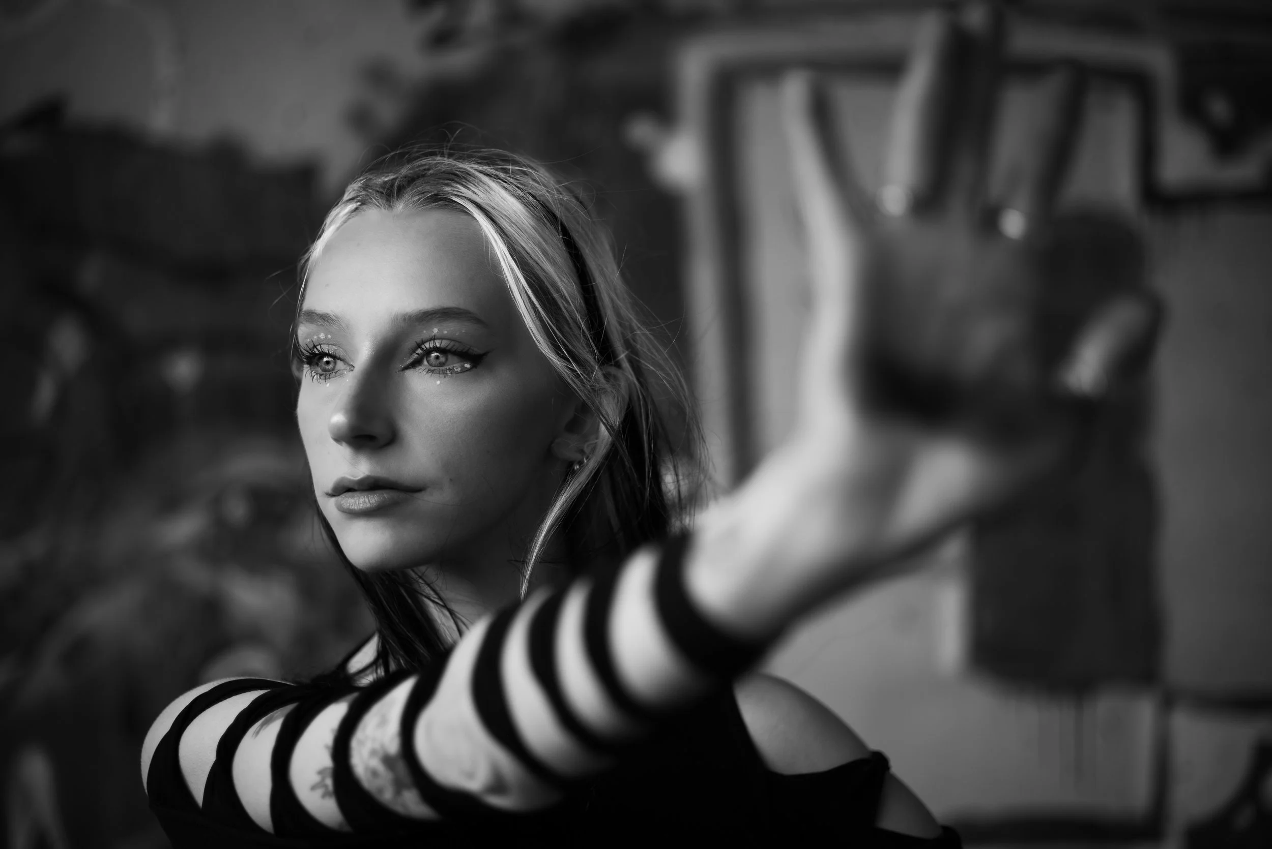A black and white photo of a woman with blonde hair and striking makeup, reaching out towards the camera with an outstretched arm wearing black and white striped sleeves, with art supplies on a wall in the background.