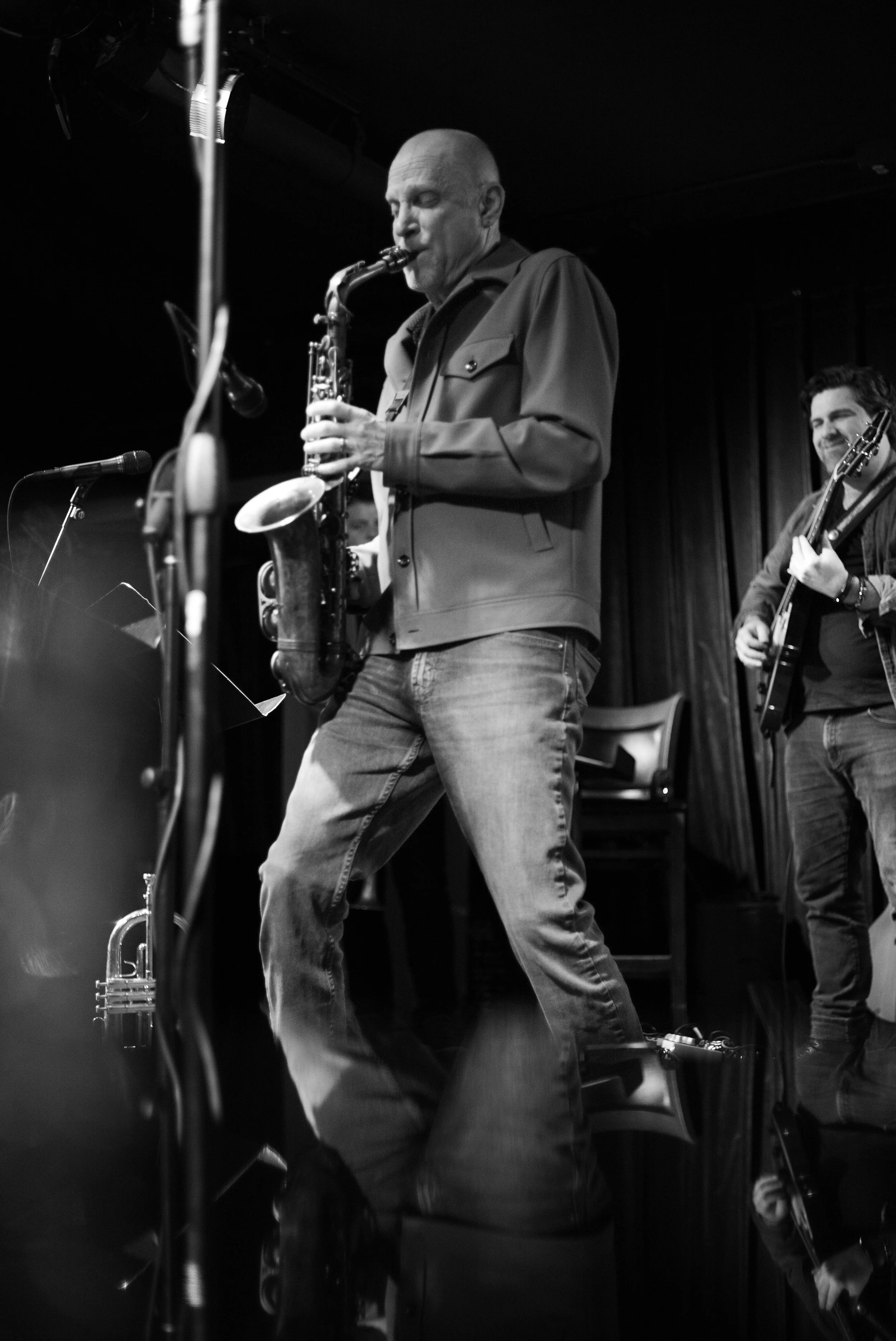 A black and white photo of a man playing a saxophone on stage, with another musician playing a guitar in the background, both smiling.