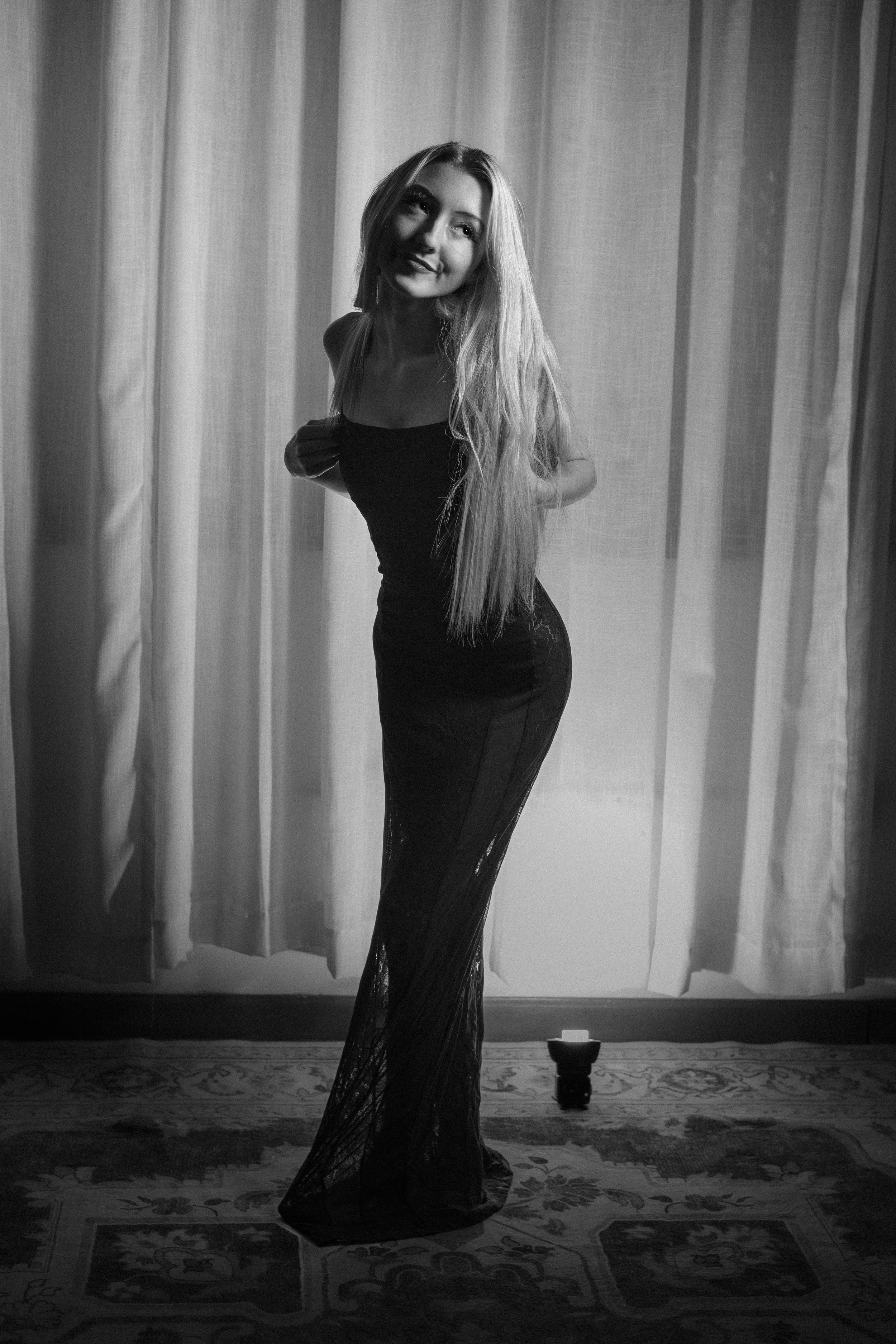A woman with long hair posing in front of curtains, wearing a fitted dress, in a black and white photo.