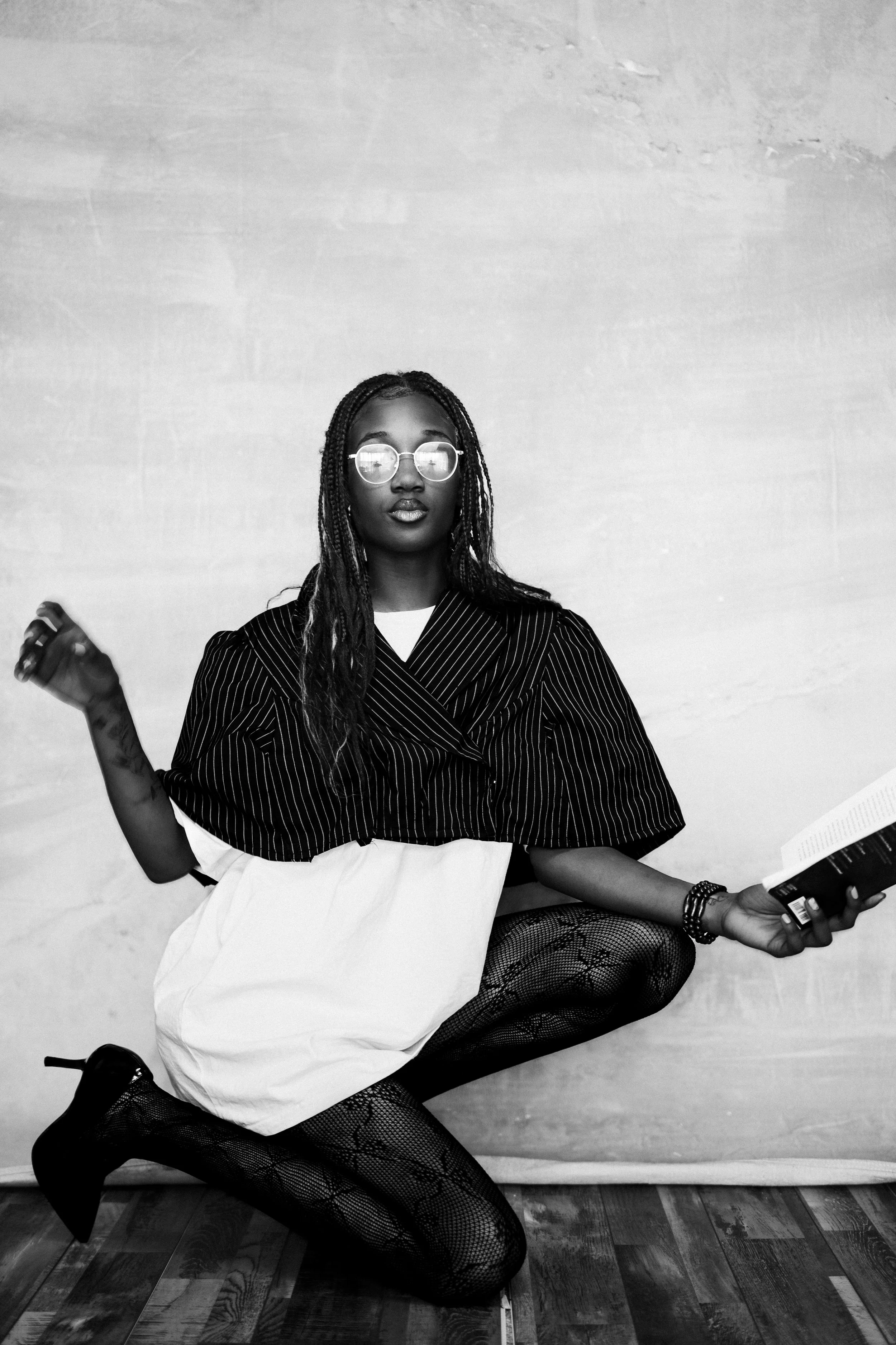 A woman with glasses and long braids, wearing a striped jacket, white skirt, patterned tights, and heels, is squatting against a plain wall while holding a book.