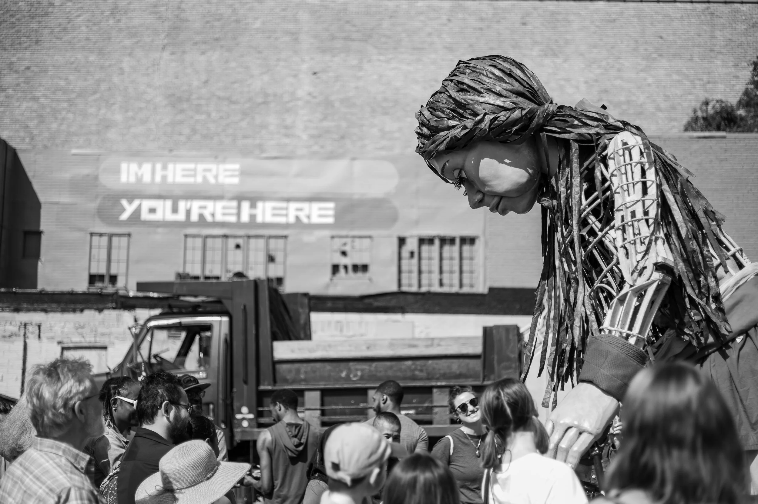 A large outdoor gathering with a diverse group of people in front of a brick building, featuring a large human-like sculpture of a girl with long, textured hair, bending forward.