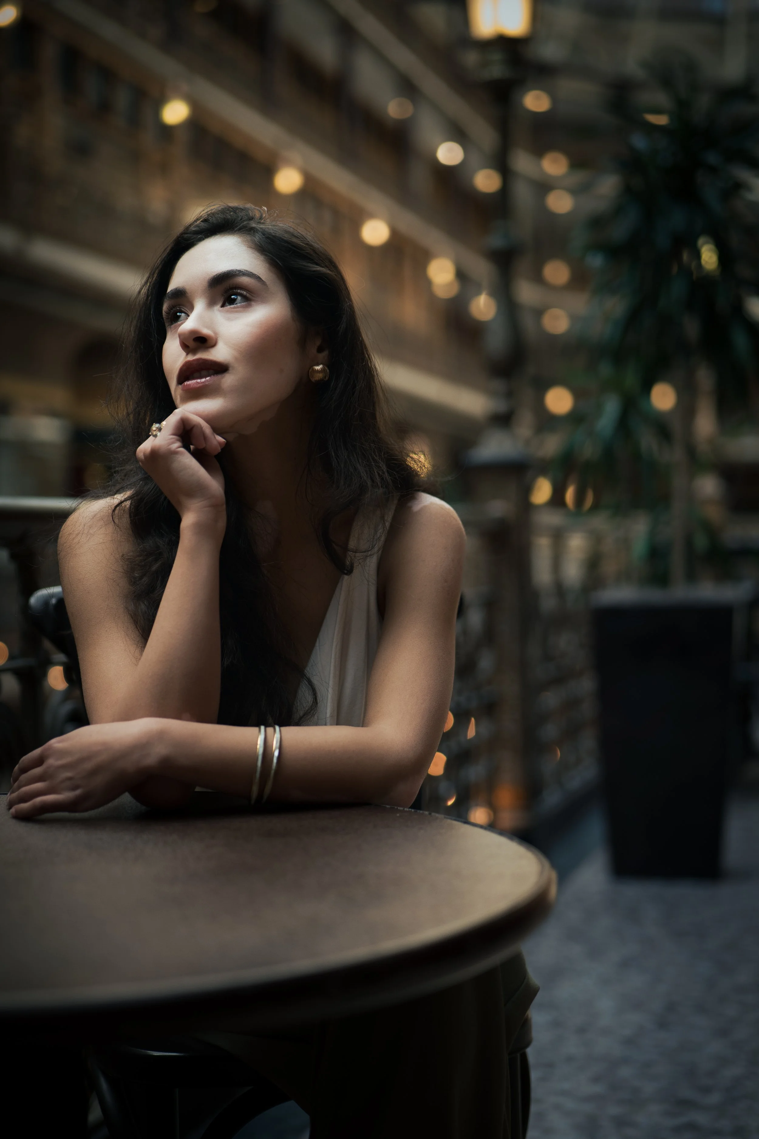 A woman with dark hair and gold jewelry sitting at a table in a dimly lit indoor space, possibly a restaurant or cafe, with warm string lights and a potted plant in the background.