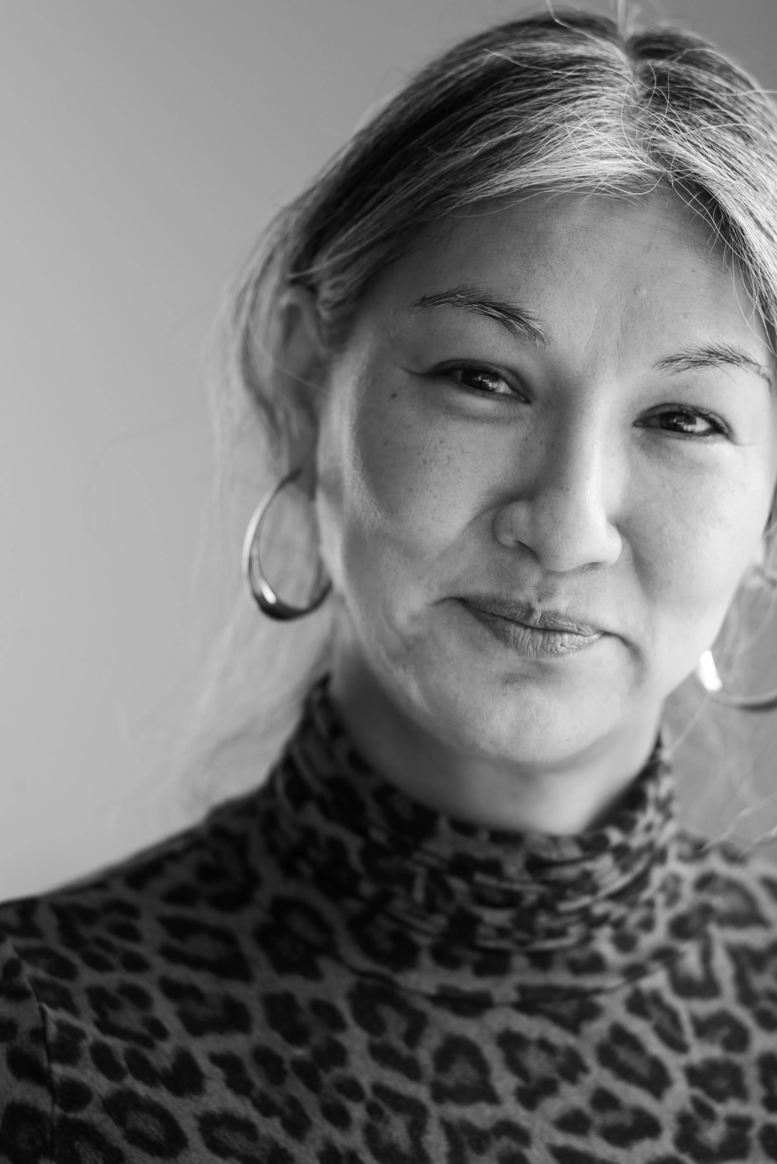 Close-up black-and-white portrait of a woman with light skin, freckles, hoop earrings, and animal print turtleneck, smiling gently.