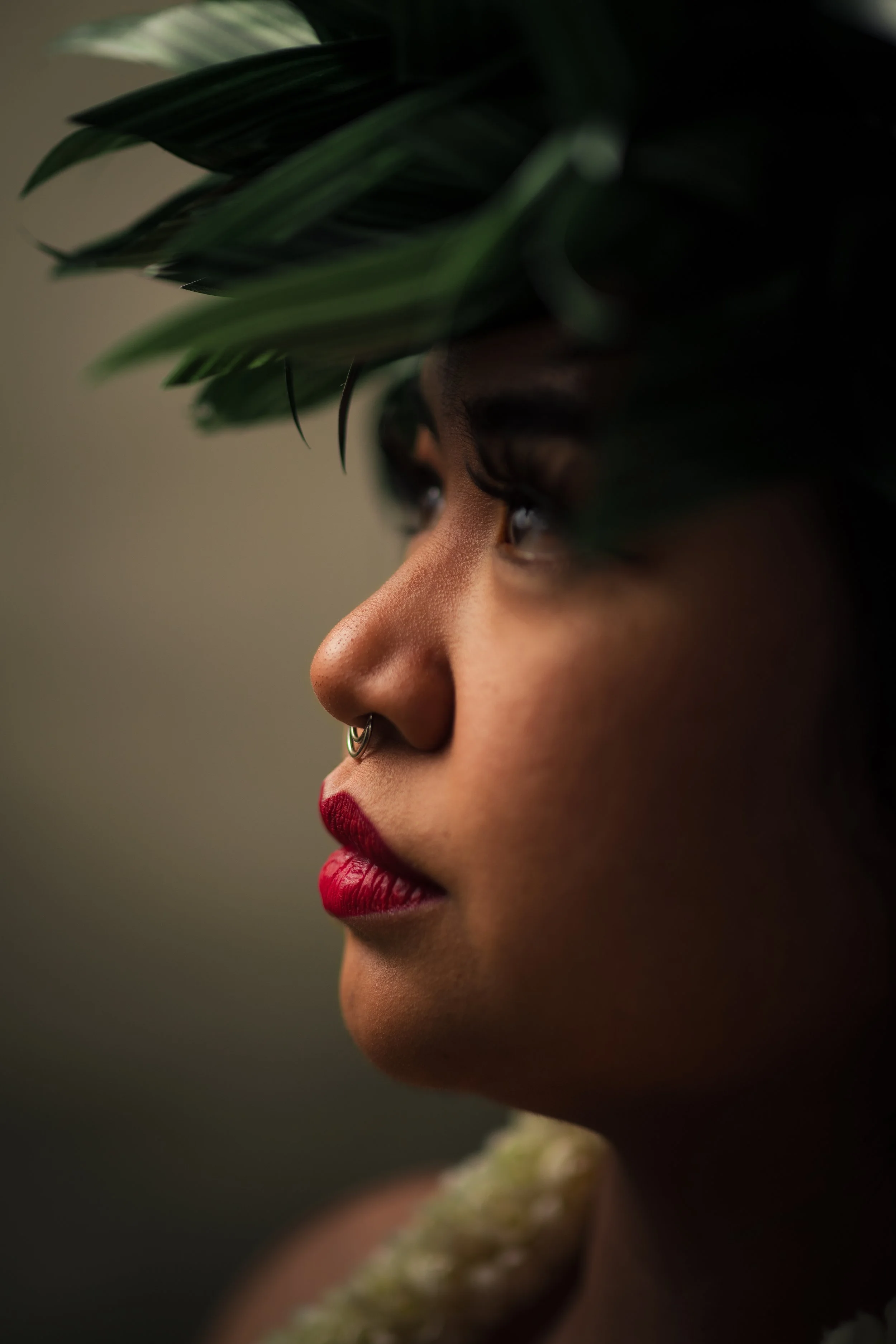 A close-up profile of a woman with medium skin tone, red lipstick, a septum piercing, dark eye makeup, wearing a leafy headpiece, and a floral necklace.