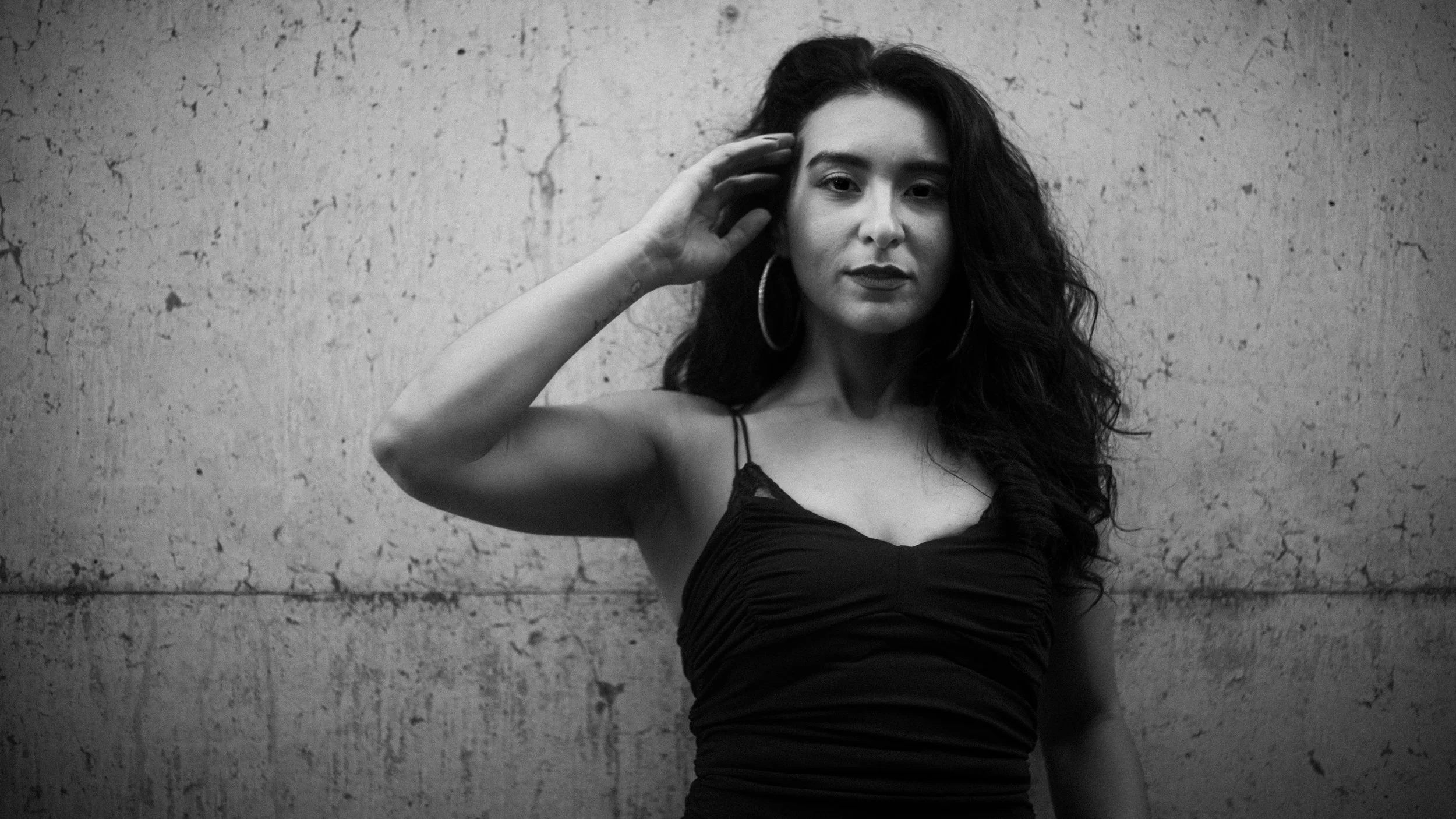 A woman with long curly hair wearing a black sleeveless top and hoop earrings, standing against a textured wall.