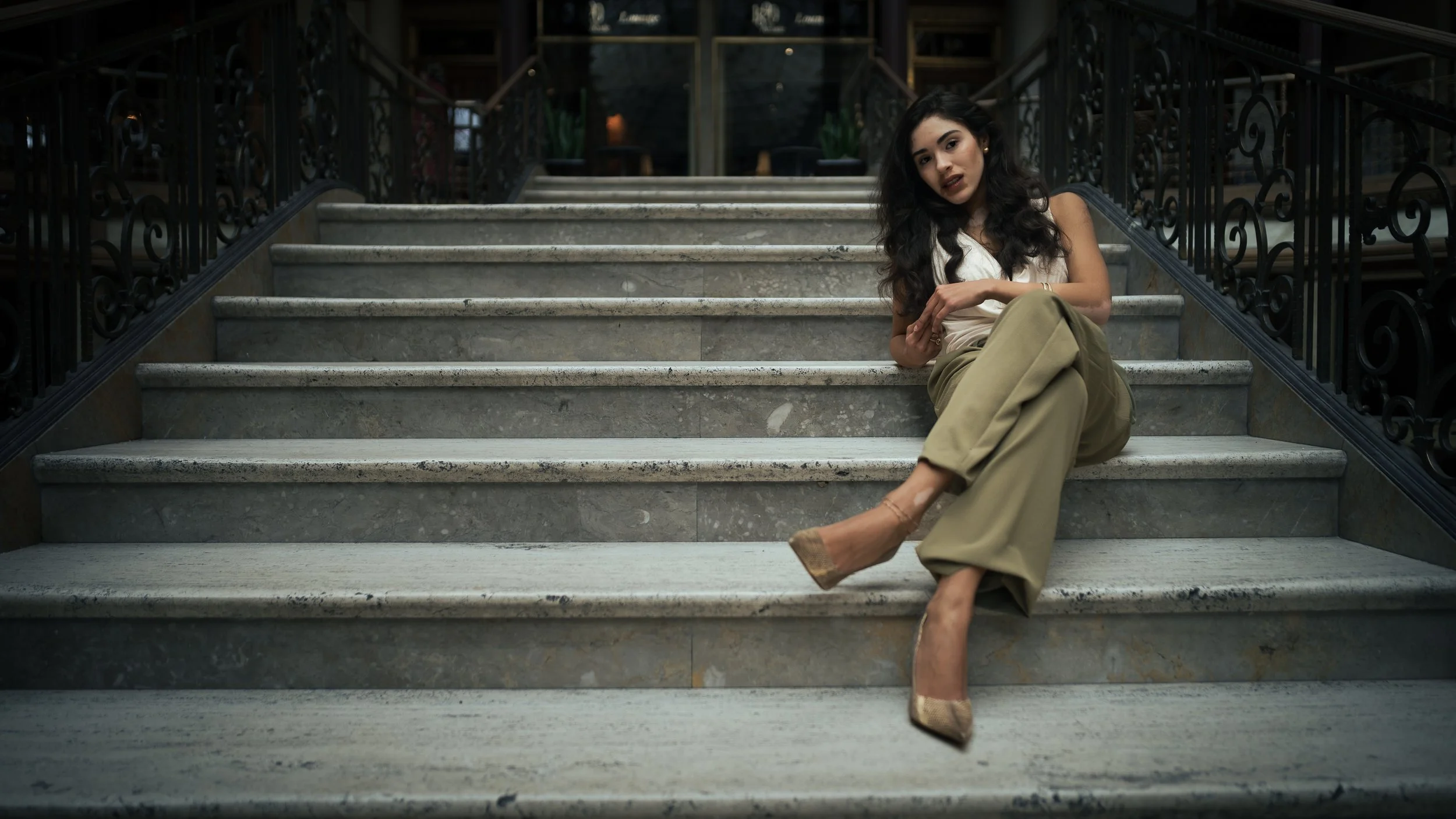 A woman with long dark hair sitting on marble stairs inside a building, wearing beige wide-leg pants and high heels, with ornate black railings on either side.