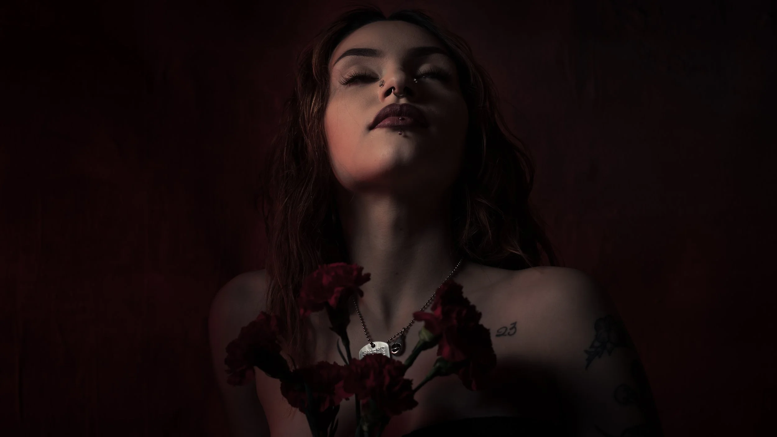 A young woman with red wavy hair and tattoos, holding red carnations, with her eyes closed, against a dark background.