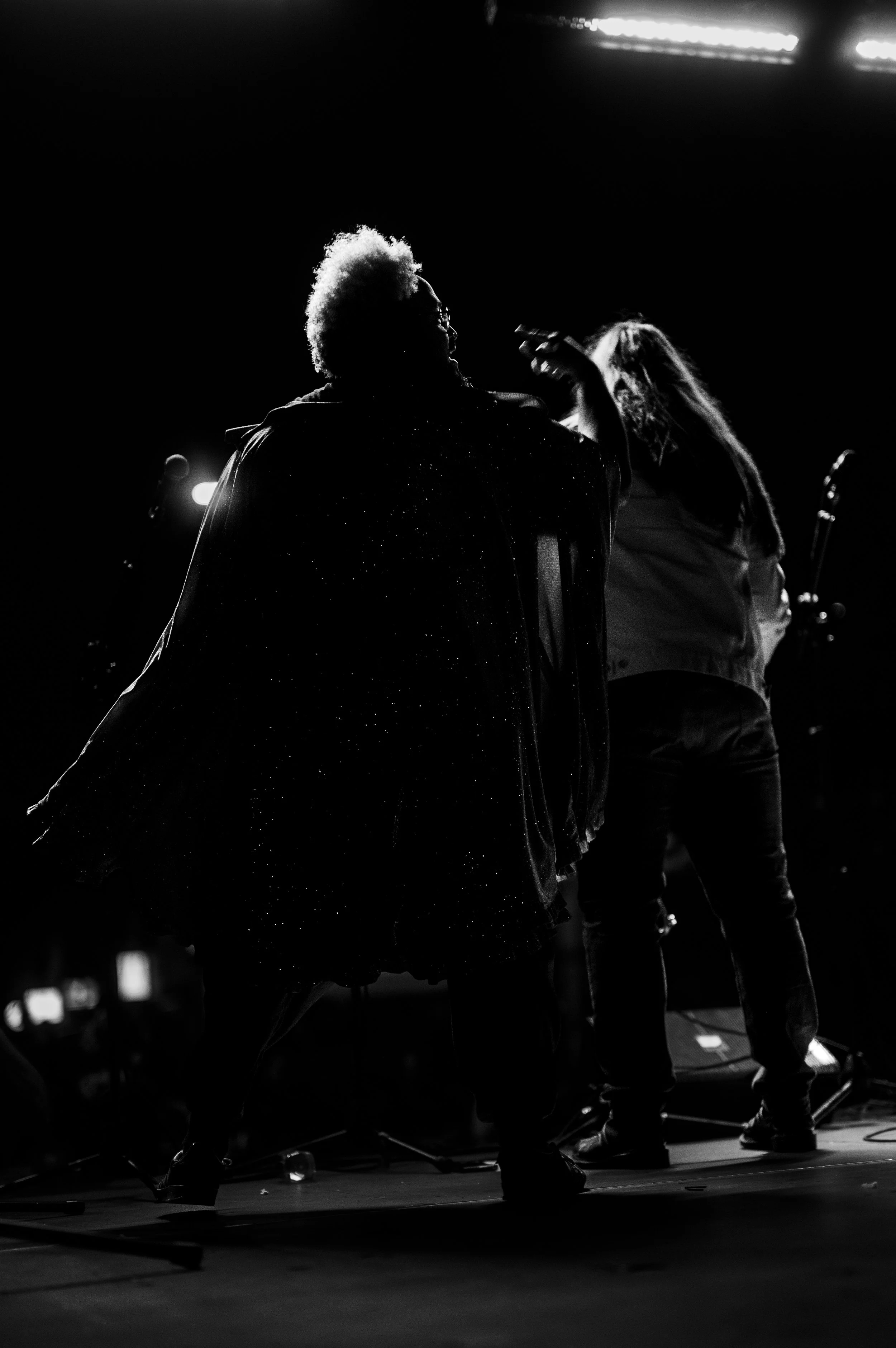 Silhouette of two performers on stage, one with an afro hairstyle, against a dark background with bright lights overhead.