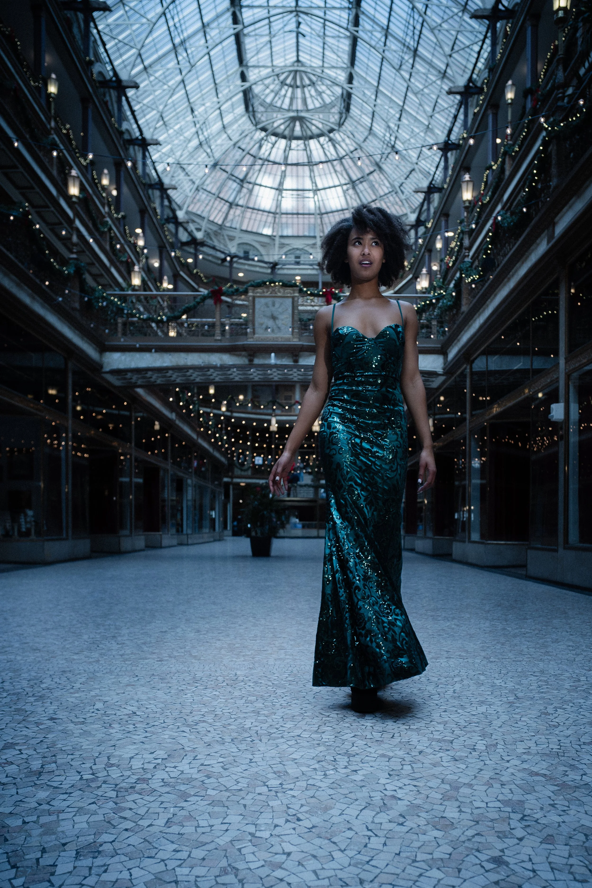 A woman in a teal, sequined evening gown walking through a mall with Christmas decorations and string lights, beneath a glass-domed ceiling.