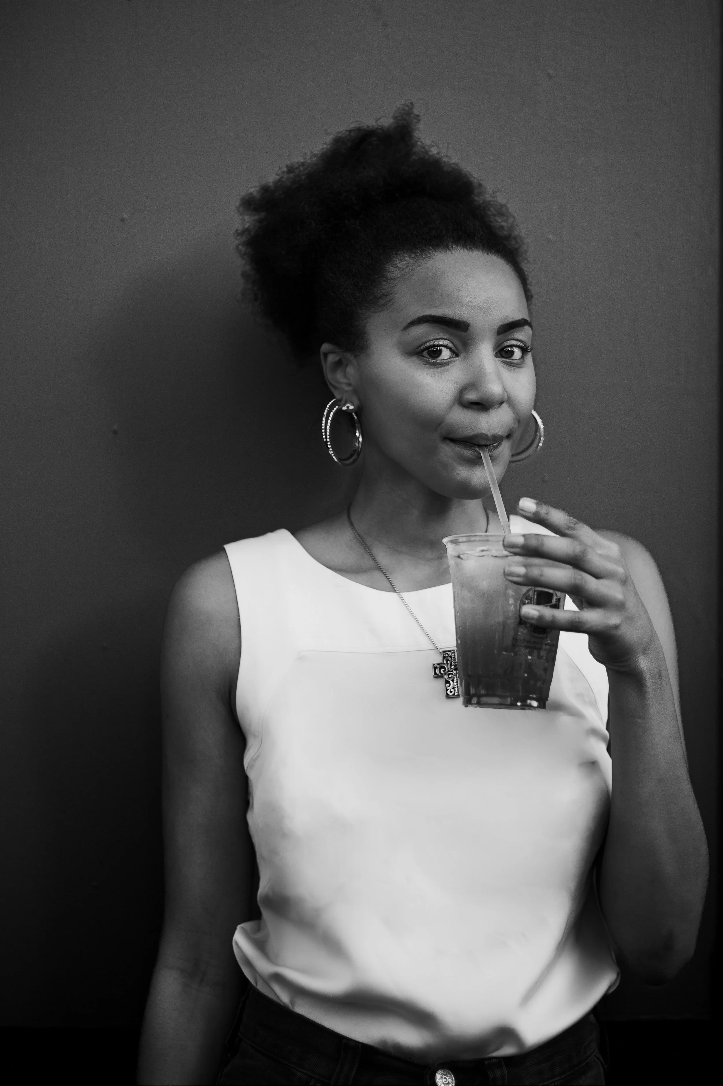 A young woman with curly hair wearing earrings and a sleeveless top, sipping a beverage through a straw from a plastic cup, standing against a plain wall.