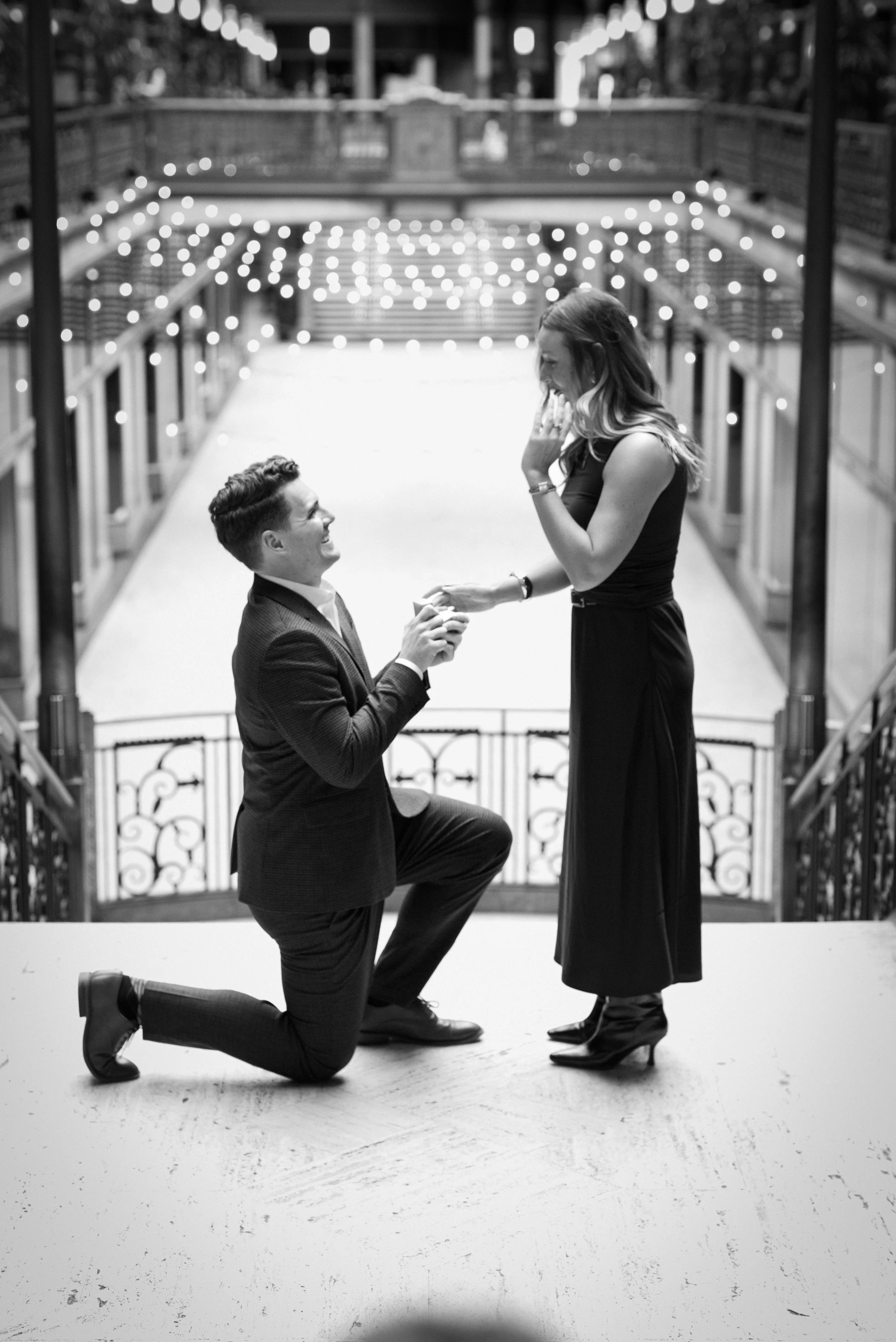 A man proposing marriage to a woman on a balcony with string lights, both dressed in formal attire, in black and white photography.