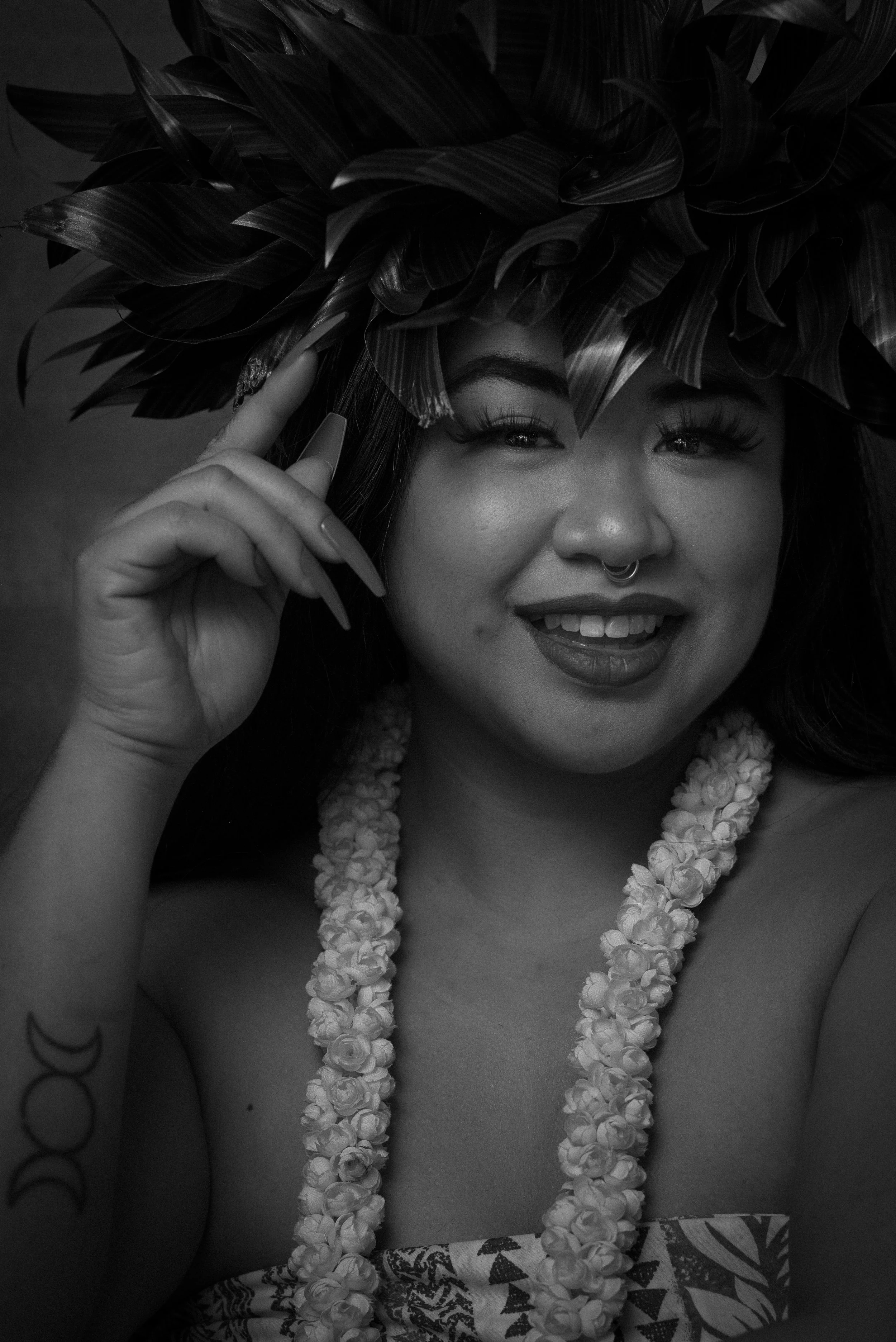 A smiling woman wearing a floral lei and a patterned dress, holding a large leafy headdress on her head, looking cheerful in a black-and-white photo.